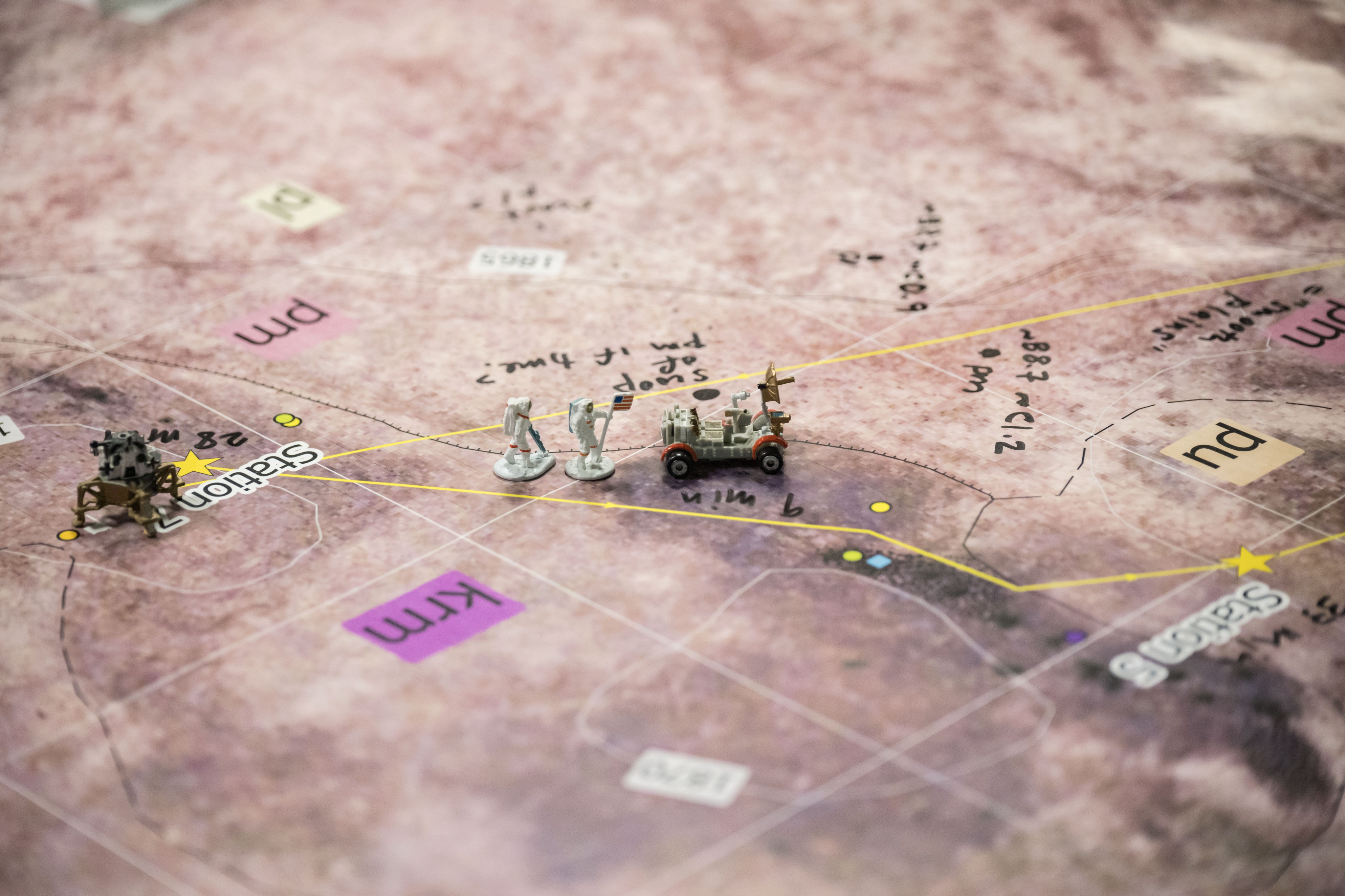 The image shows a closeup of a map that’s pinkish in color with small, shaded areas. There are labels on the map, such as “krm” and “pu,” and dotted lines, small dots and squares and stars that mark locations on the map. A miniature lander model, smaller than the palm of the hand, is sitting at a location on the map labeled “Station 7.” Two miniature astronaut figures, one holding a U.S. flag, are standing a few inches to the right of the lander, and to the right of them sits a miniature rover.