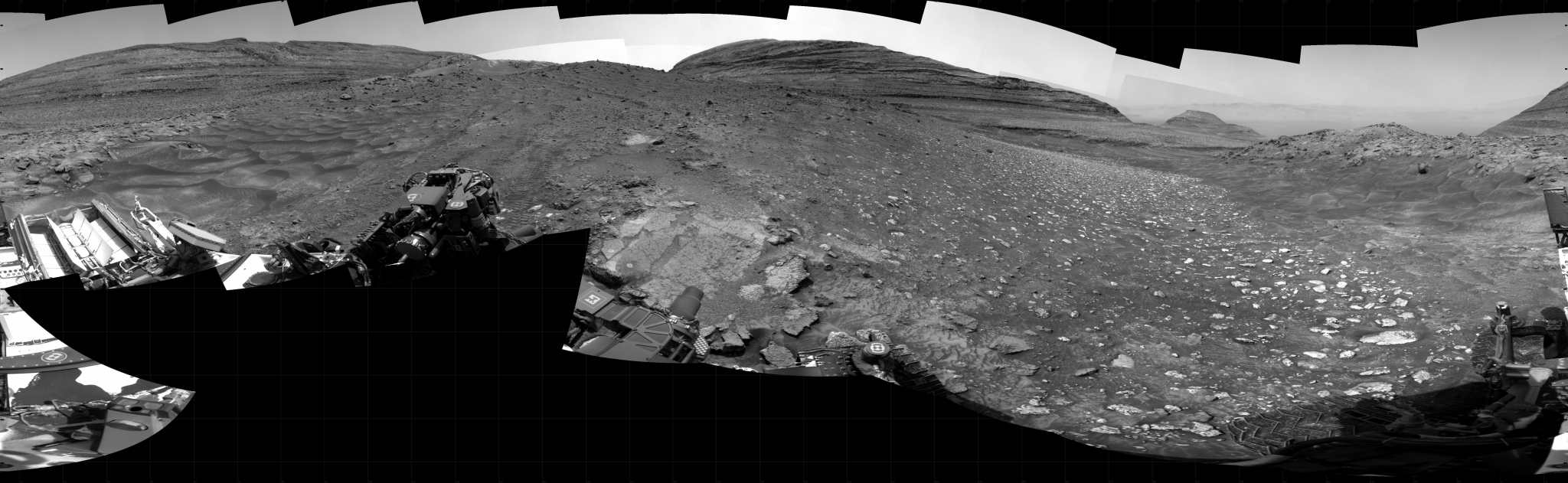 A black and white photographic panorama of the surface of Mars, showing rolling hills in the distance, with the foreground a mixture of areas with soil, gravel and small rocks, of windswept dunes, and of large broken slabs of flat rock.