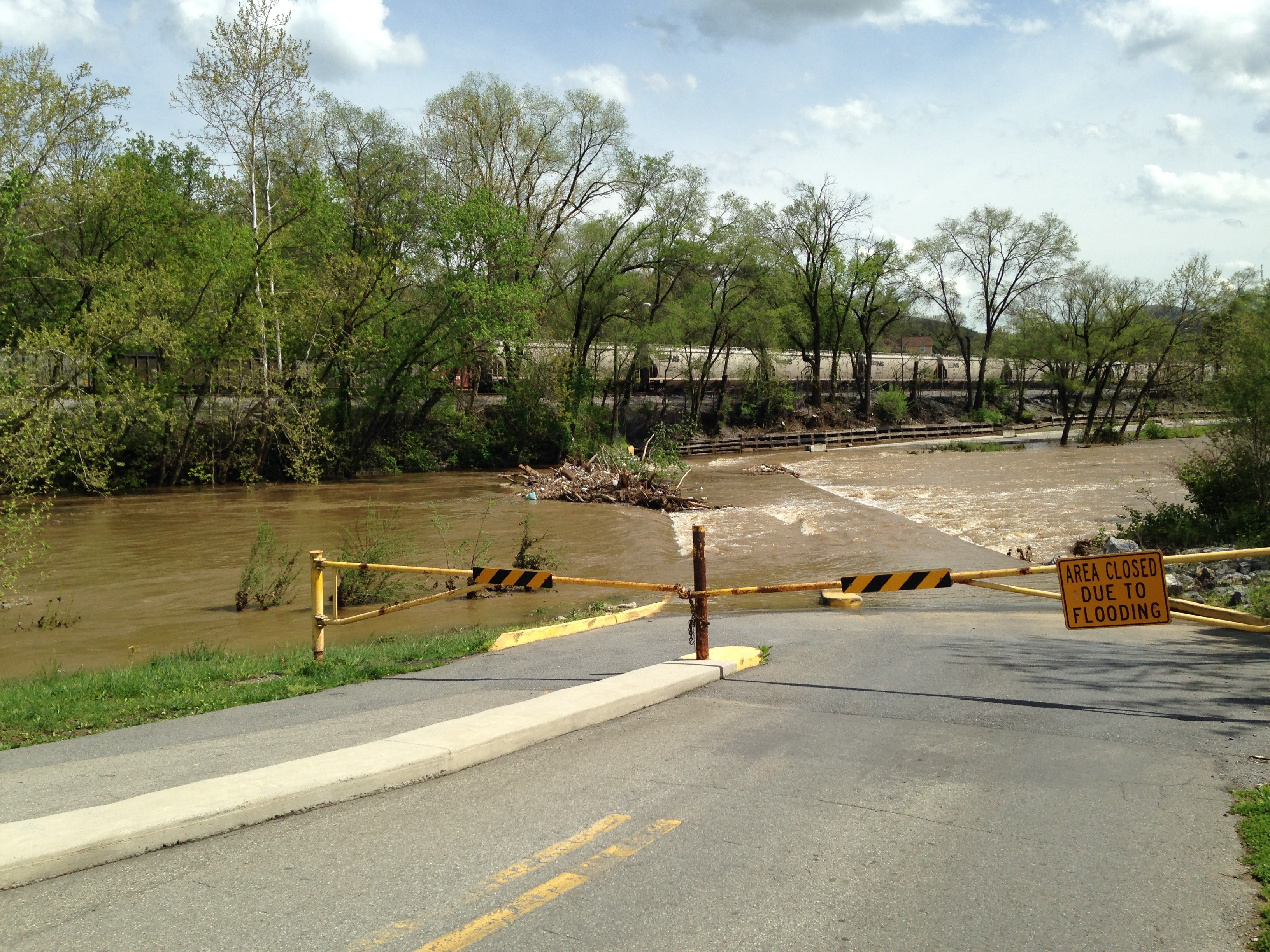 The image shows a road leading to a river or stream that is currently experiencing flooding. The road is blocked off with a yellow and black striped barrier and a sign that reads "AREA CLOSED DUE TO FLOODING." The water level in the river is high, and the current appears strong, with debris visible in the water. The surrounding area is lush with green trees and vegetation, indicating a rural or natural setting. The sky above is partly cloudy.