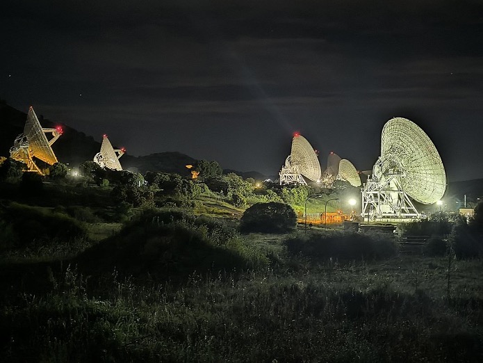 A nighttime photograph of a field with several large satellite dishes positioned across the landscape. The dishes are illuminated by lights, creating a stark contrast against the dark sky.
