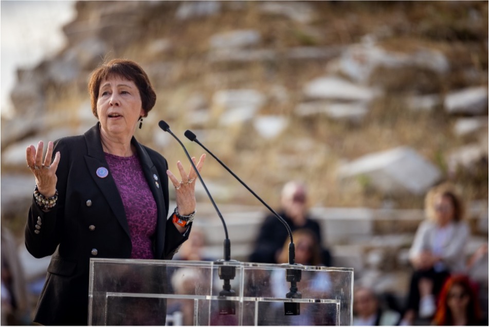A woman giving a speech outdoors, standing at a clear podium with two microphones. She is wearing a black blazer with a purple shirt and gesturing with her hands. There are blurred people and rocks in the background.