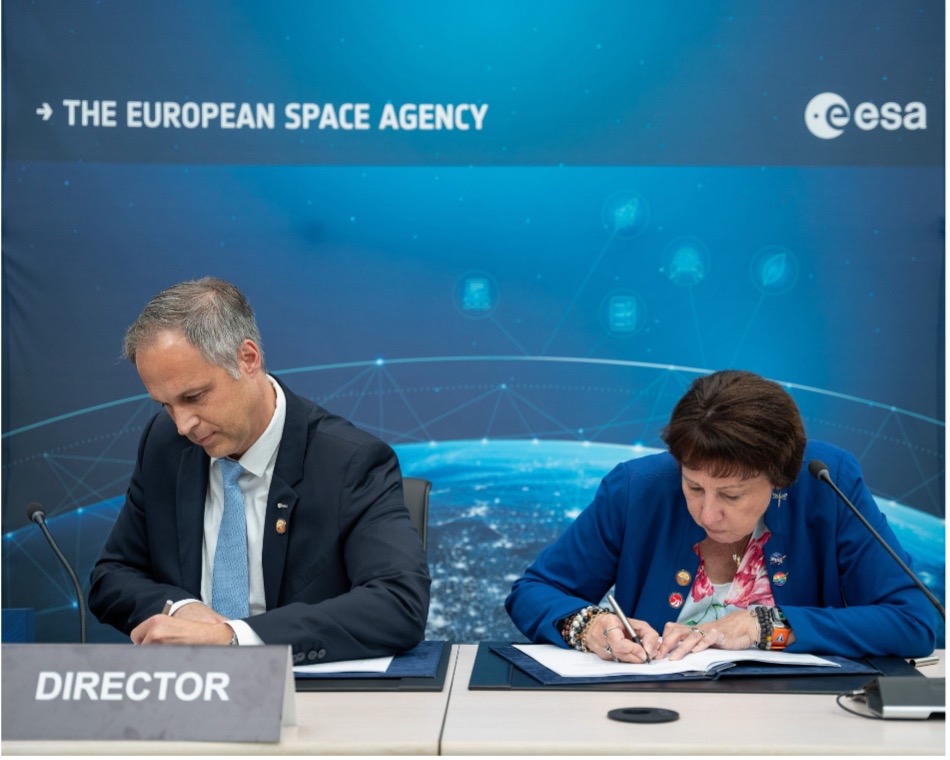 A man and a woman sitting at a table, both signing documents. The background has a banner reading "The European Space Agency (ESA)" with a blue, space-themed design. The woman is on the right, wearing a blue blazer, and the man is on the left, wearing a dark suit and light blue tie.