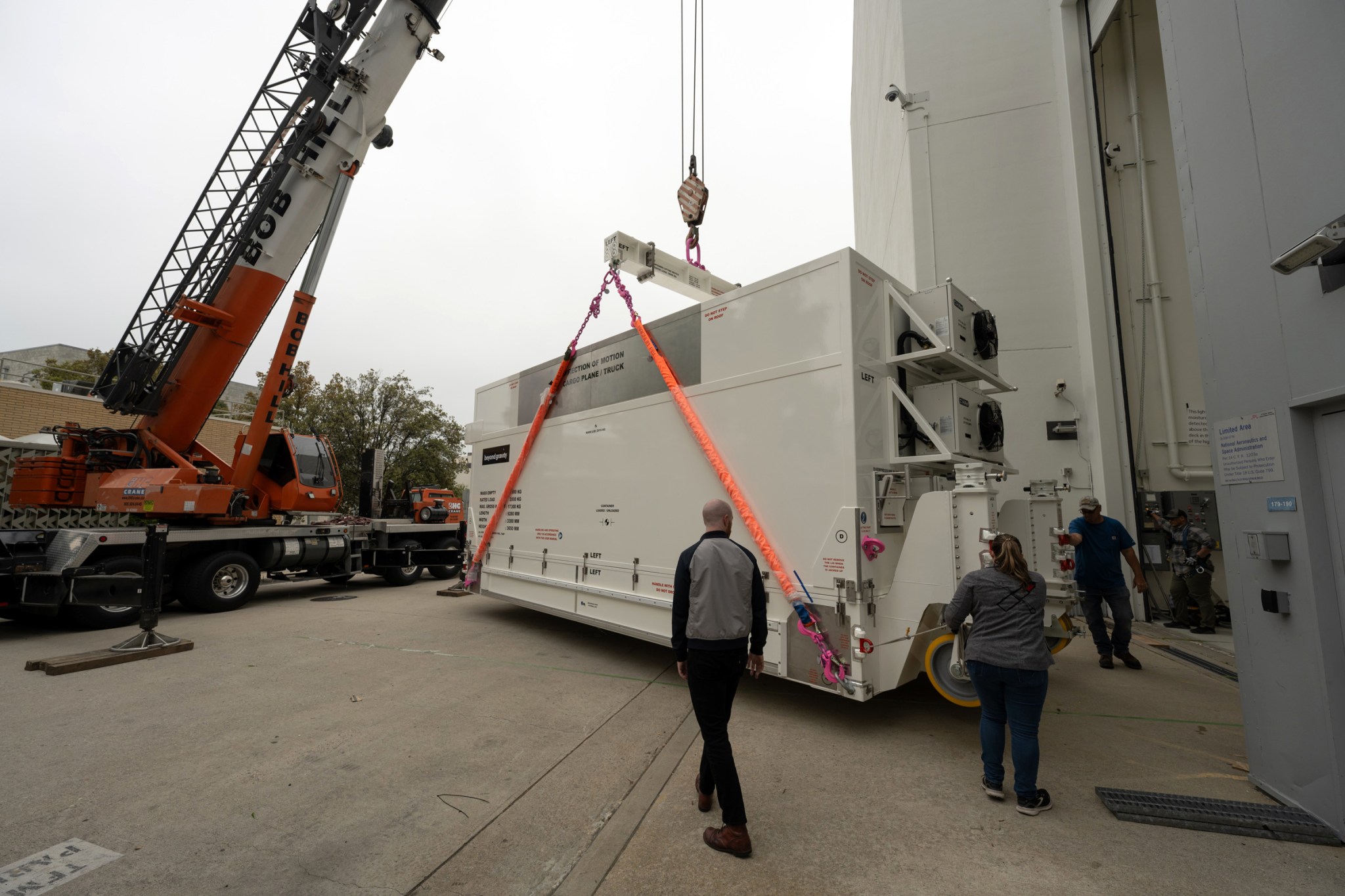 Workers stand nearby as a large white container with spacecraft equipment is lowered to the ground by a crane at NASA's Jet Propulsion Laboratory.