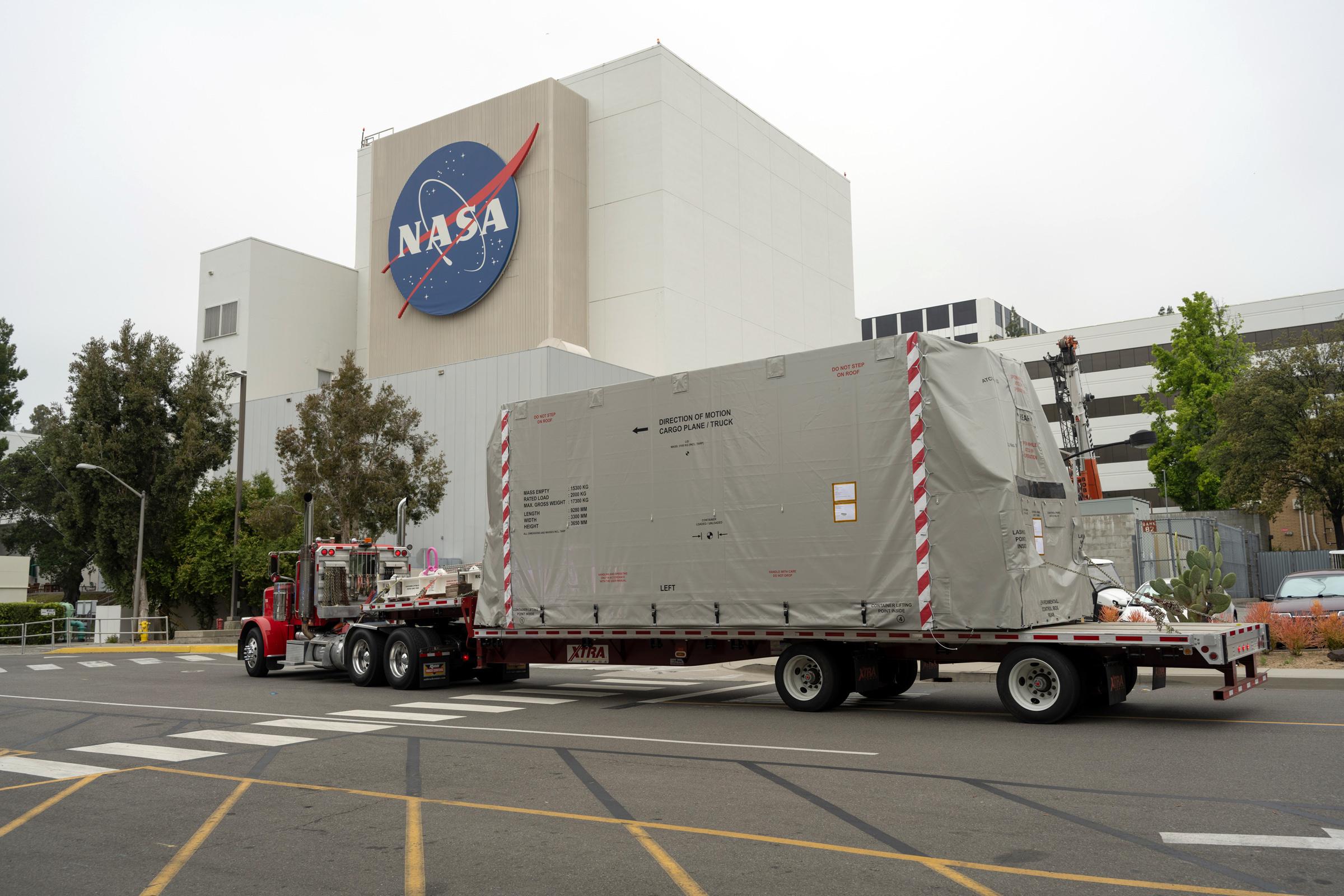 A truck arrives in front of a large white building at the Jet Propulsion Laboratory with the NASA logo on it.