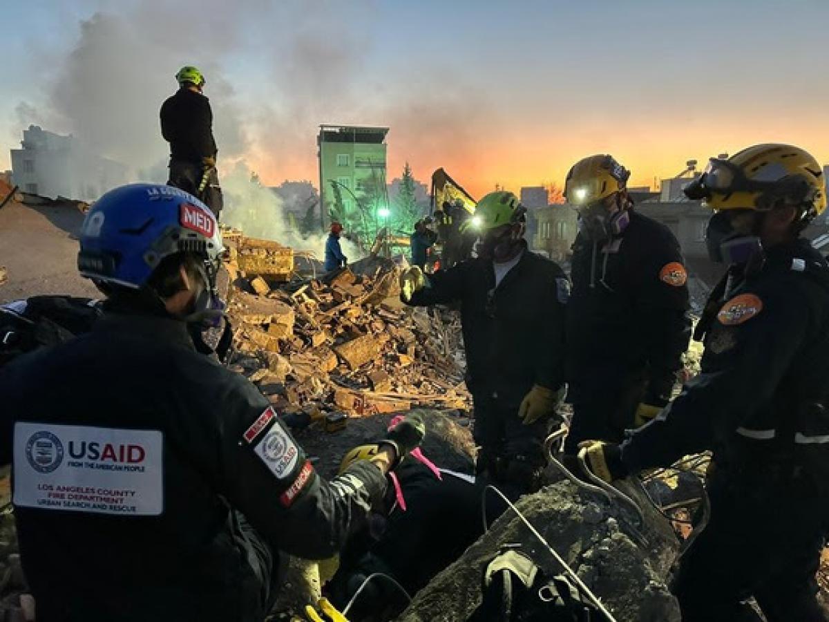 people in search-and-rescue attire stand among rubble