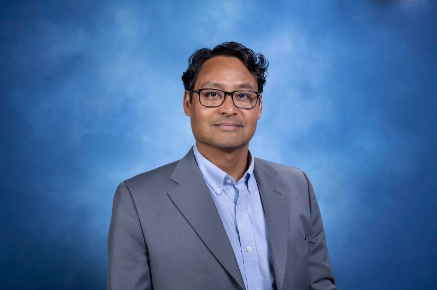 Headshot of Rahul Ramachandran in front of a blue background. He wears a light blue shirt, dark gray jacket, and glasses.