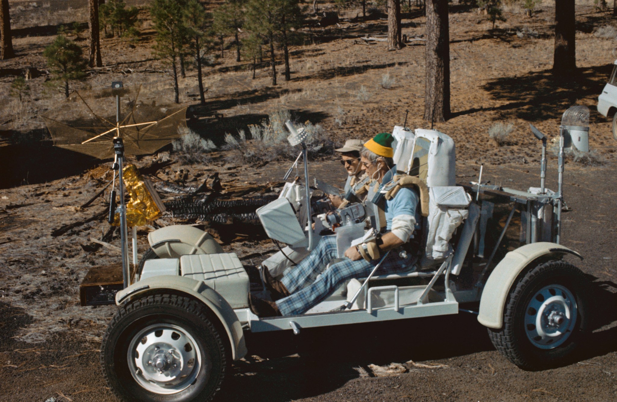 Two people are sitting in a large vehicle with no roof, strapped into large, rectangular seats. The vehicle is sitting on brown soil. Spruce trees are in the background. The two people are looking at a box in front of them. Antennas stretch up from different parts of the vehicle.