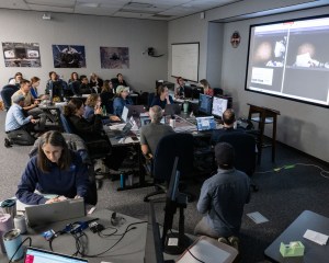 People are sitting in a typical, brightly lit office-building room. Colorful posters line the walls. A large screen is in the top right corner, showing two side-by-side images of shapes that are hard to make out. People are sitting around tables, some are kneeling, looking either at the large screen or at the small computer screens in front of them.