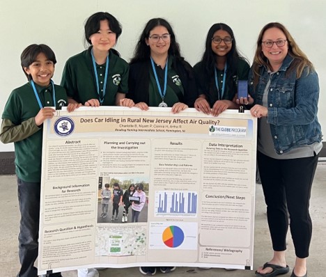 Four students and their teacher smiling, holding their research poster titled "Does Car Idling in rural New Jersey affect air quality?" The students have medals.
