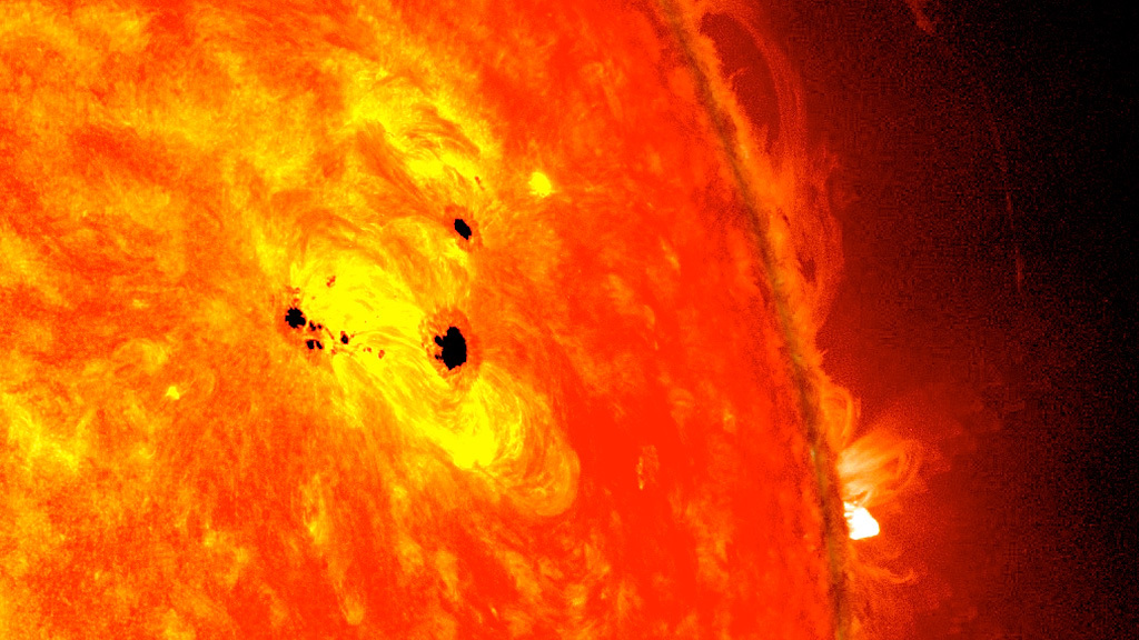 A portion of the Sun, shown in dark orange and yellow. Over one bright yellow region are a few splotches of black – sunspots.