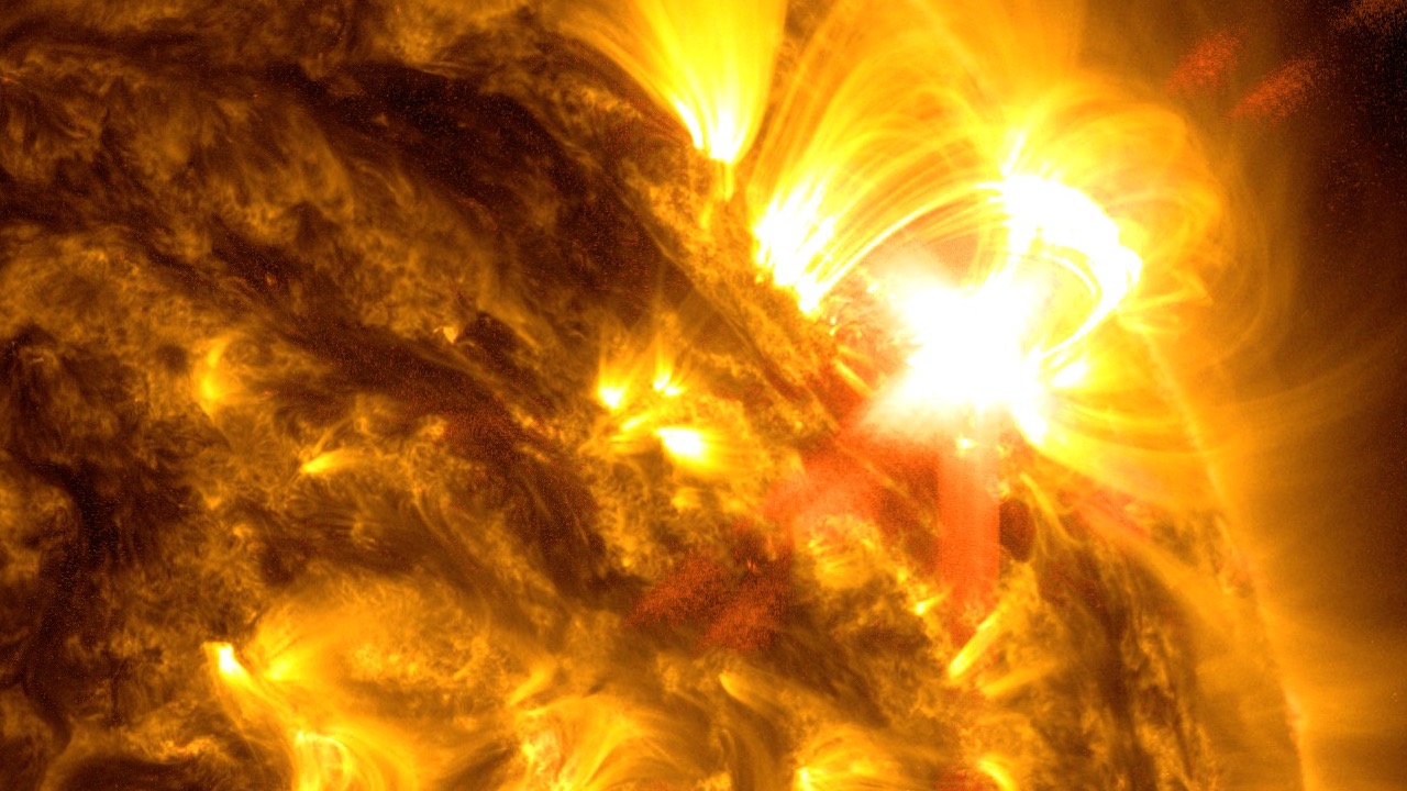 A close-up portion of the Sun appears in shades of yellow, brown, and white. A bright white burst of light, a solar flare, appears in the upper right.