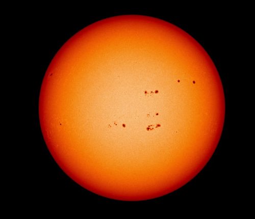 An image of the Sun. The Sun is yellow and orange and speckled with several dark splotches – sunspots.