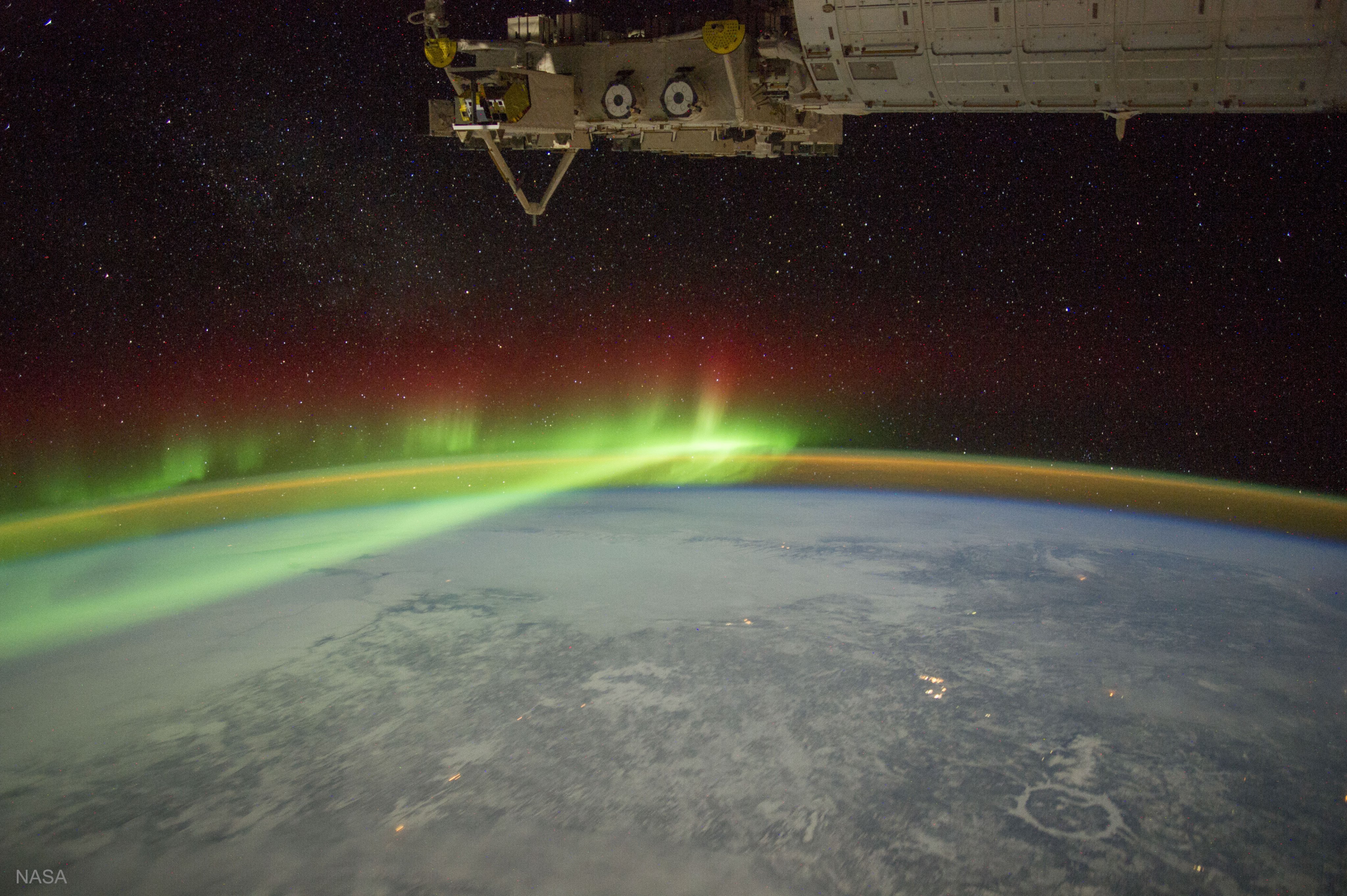 Image of Earth at night with lights glowing on Earth’s surface. Above Earth are green, and red bands of light signifying airglow. Above the bands of light is a black space with white stars and an orange and green band of light below the airglow.