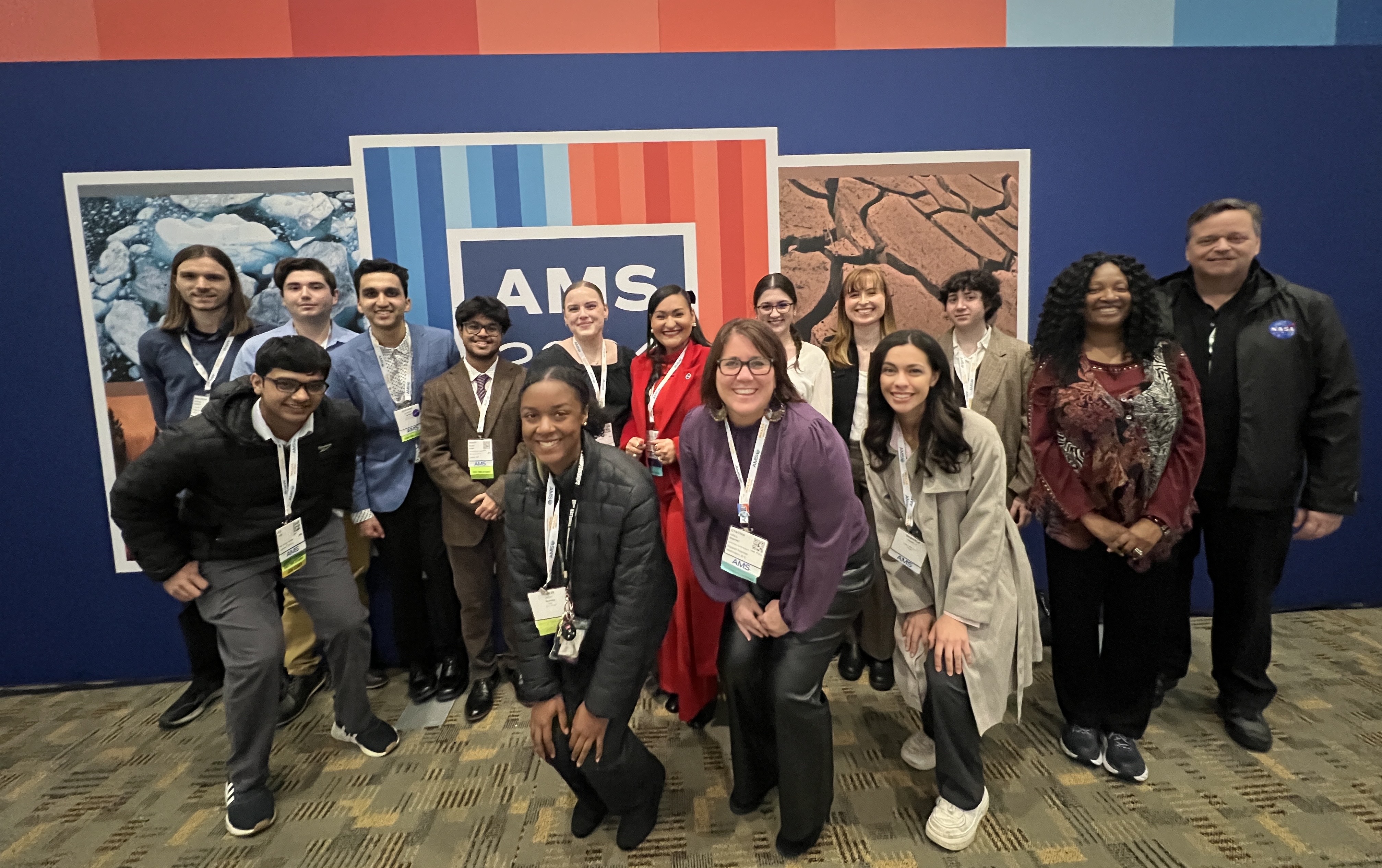 15 people gather for a photo in front of a blue wall with AMS 2024 logo over vertical striping of blue colors left and red colors right. Four people stand in the foreground to fit in the photo.