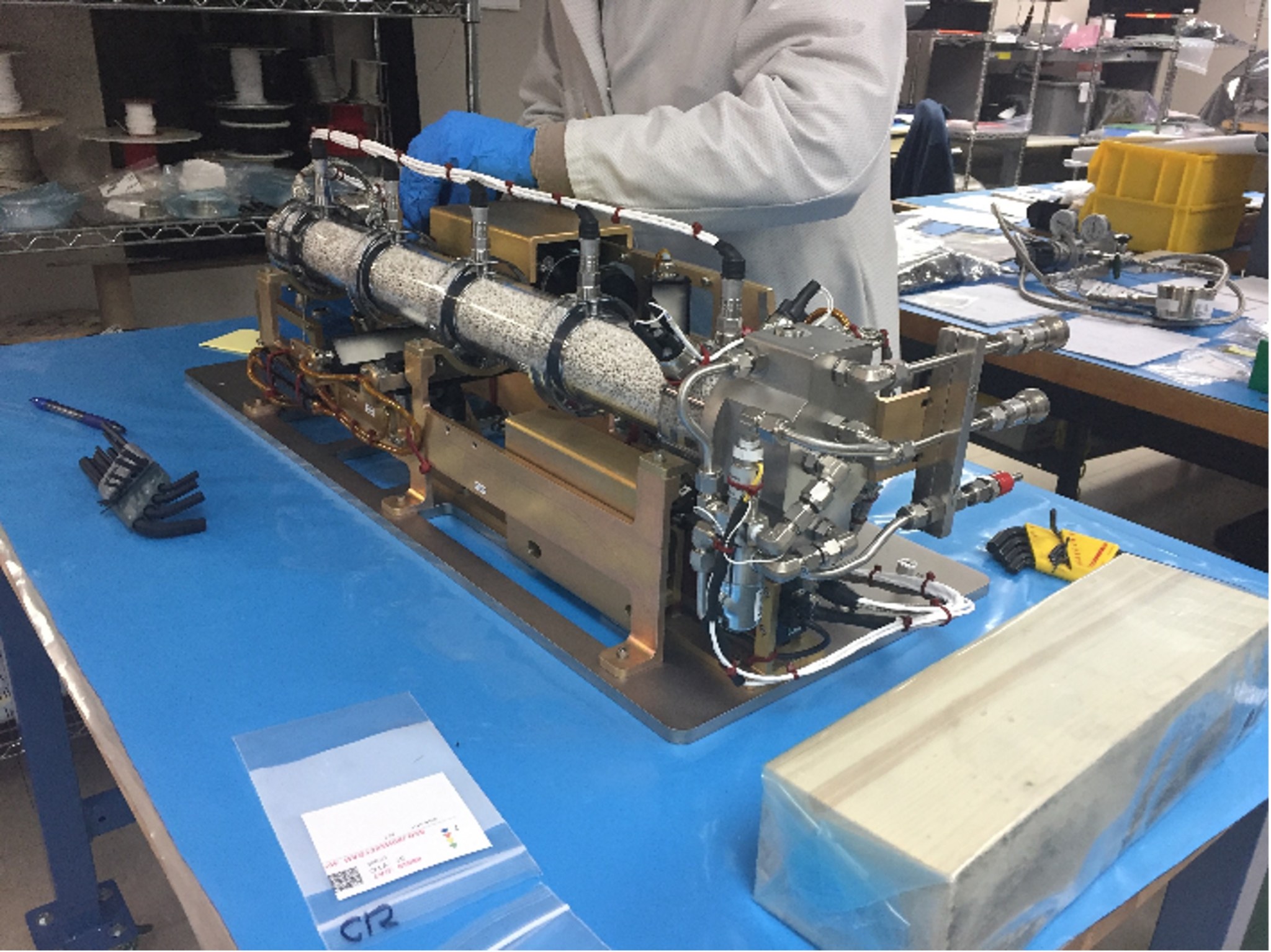 A complex scientific instrument sits atop a table lined with blue paper. The instrument is comprised of a metal framework with a grayish tube running its length. The torso of a scientist is in the background, his blue-gloved hand inspecting the instrument.