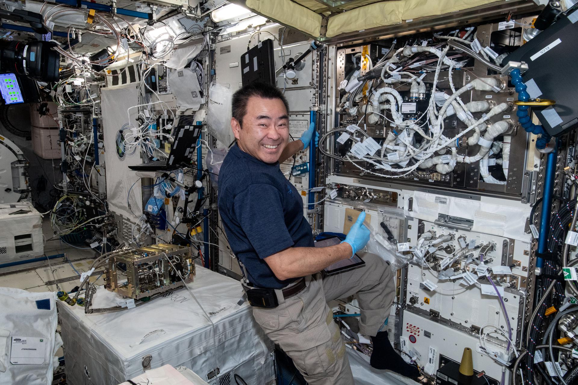 A Japanese astronaut smiles at the camera as he installs equipment against a wall of wires and hardware.