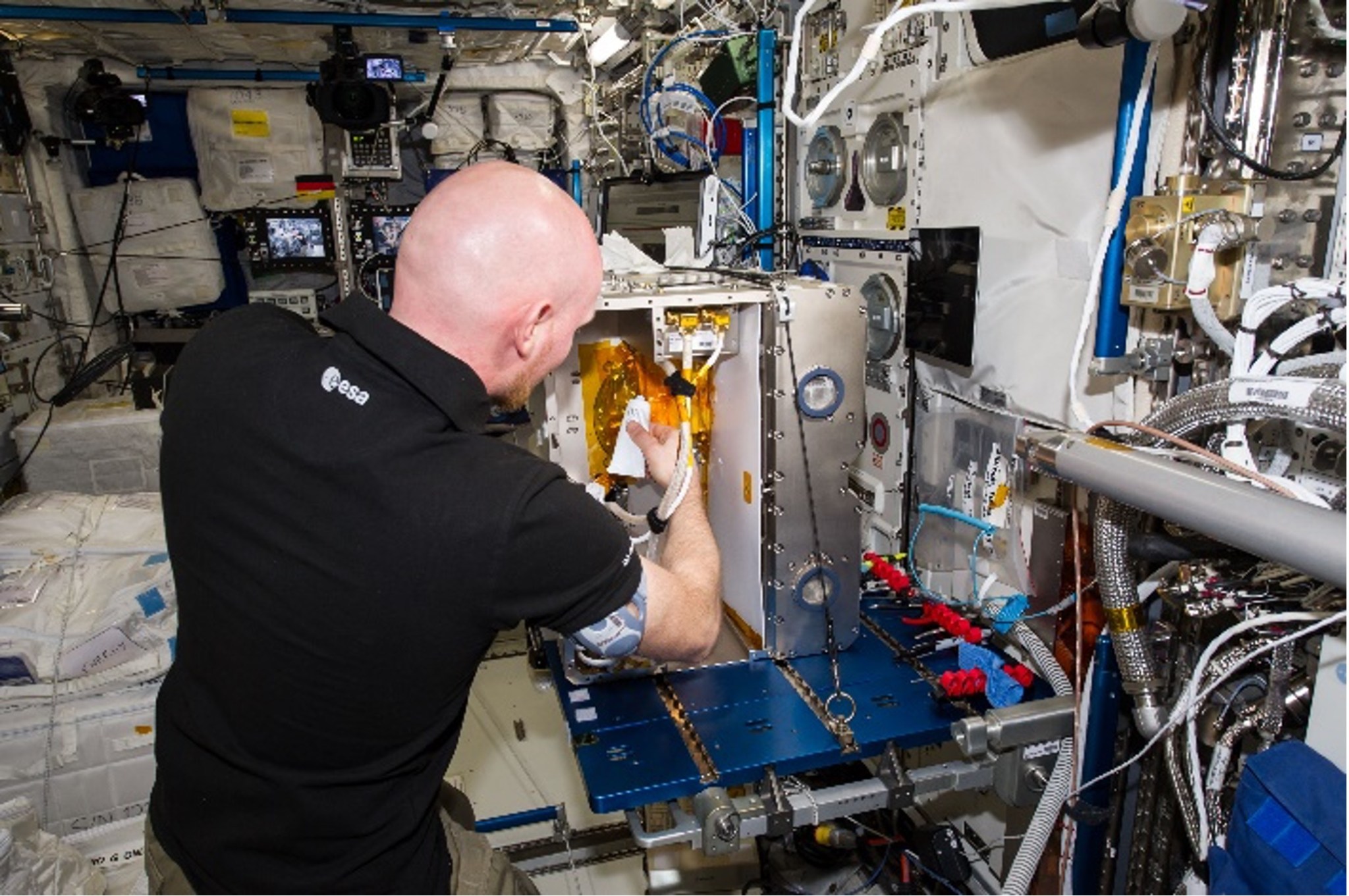 An astronaut wearing a short-sleeved black polo shirt with his back to the camera appears to be working on a piece of metal equipment, The equipment is shaped rectangular-shaped and juts out from a wall of exposed wires and other metal parts.