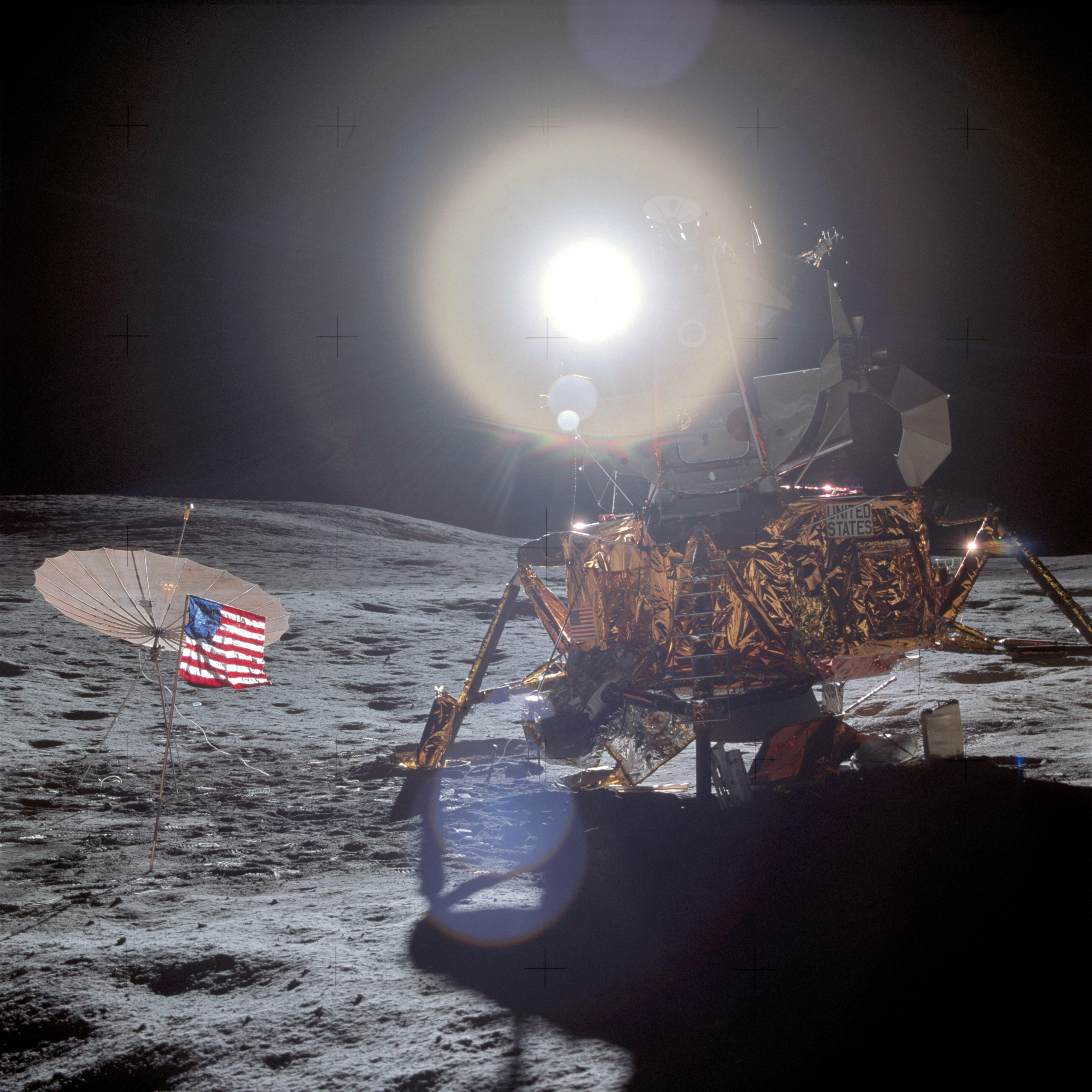 The Apollo 14 Lunar Module on the surface of the Moon. A small American flag and small antenna are planted a few feet away. In the background, the Sun creates a bright flare.