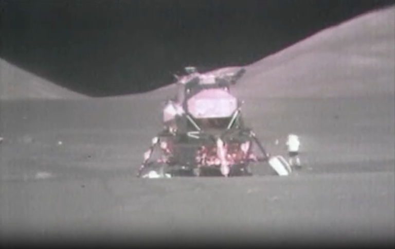 An image of the Moon lander on the surface of the gray Moon. An astronaut is small in comparison, standing on the surface.