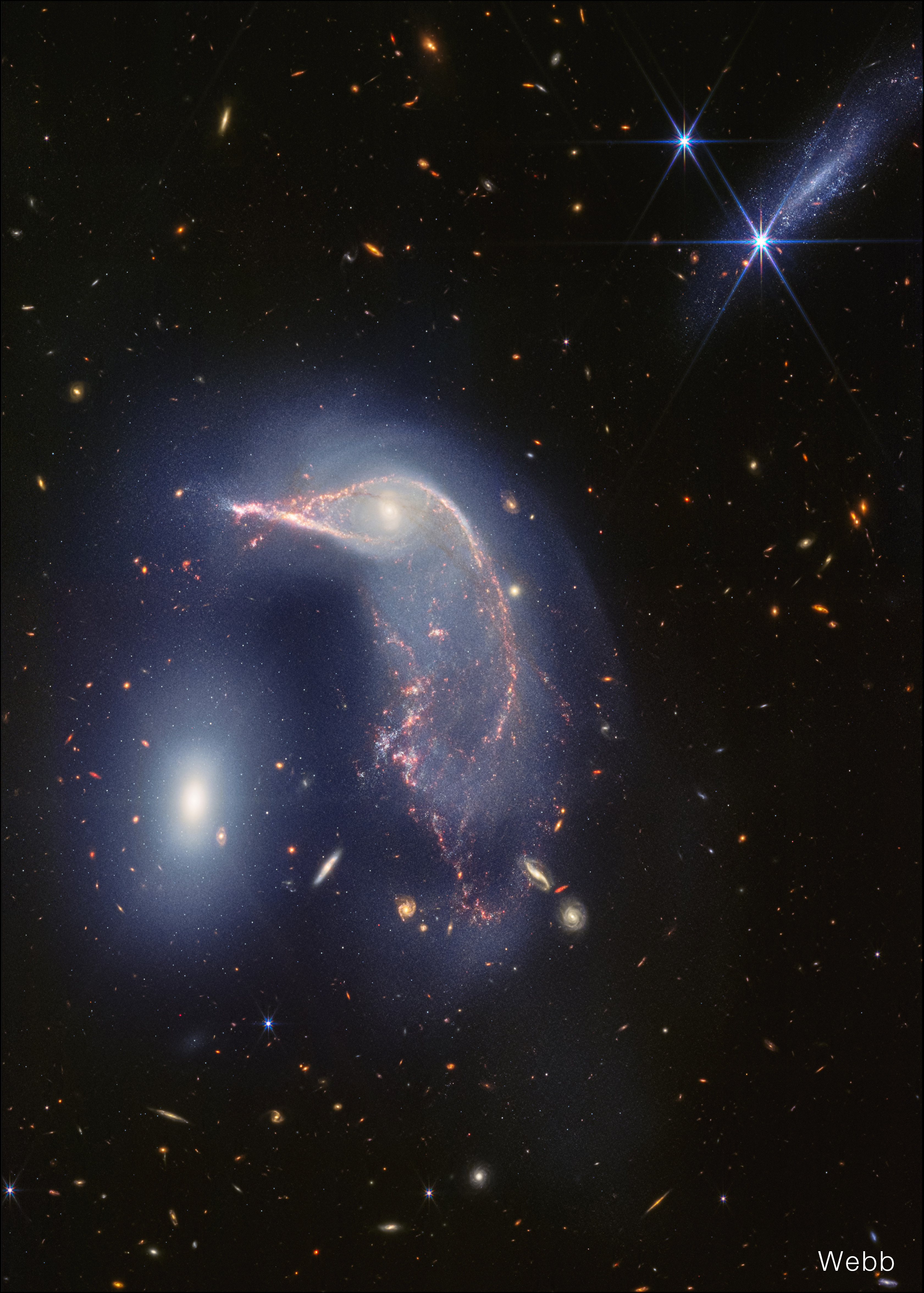Webb's view of a Frame split down the middle: Hubble’s visible light image at left, and Webb’s near-infrared image at right. Both show the Egg at left and the Penguin at right. In Hubble’s view, the Penguin is highly detailed, with a bright blue beak, body, and tail that is covered in an arc of bright brown dust. The Egg, to its left, appears bright, gleaming yellowish white. At top right is another galaxy seen from the side, about as long as the Egg’s height. Dozens of galaxies and stars appear in the background. Webb’s near-infrared image shows the Penguin’s beak, head, and back in shades of pink. It’s tail-like region is more diffuse, and a mix of lighter pinks and blues. The Egg appears slightly larger in blue layers. A semi-transparent blue forms an upside down U overtop both galaxies. At top right, an edge-on galaxy has many more pinpricks of light, which are stars. Thousands of galaxies and stars appear in the background. Some galaxies are shades of orange, while others are white. Click View Description for additional details.
