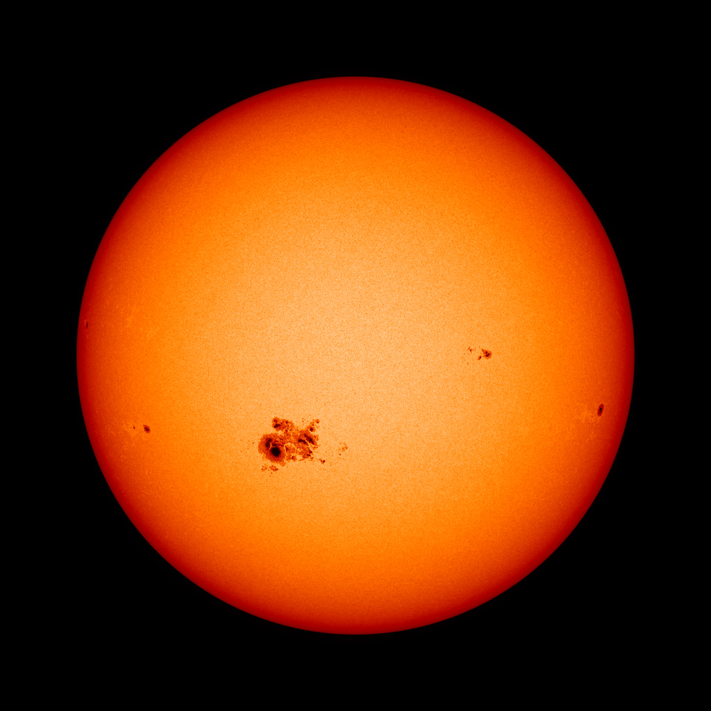 The Sun against a black background. The Sun appears mostly orange and fades to a darker red on the edges. Toward the middle and slightly to the left on the solar surface a few dark splotches.