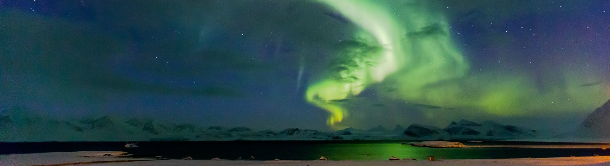 A ribbon of green aurora appears in the night sky over snowy mountains and a body of water.