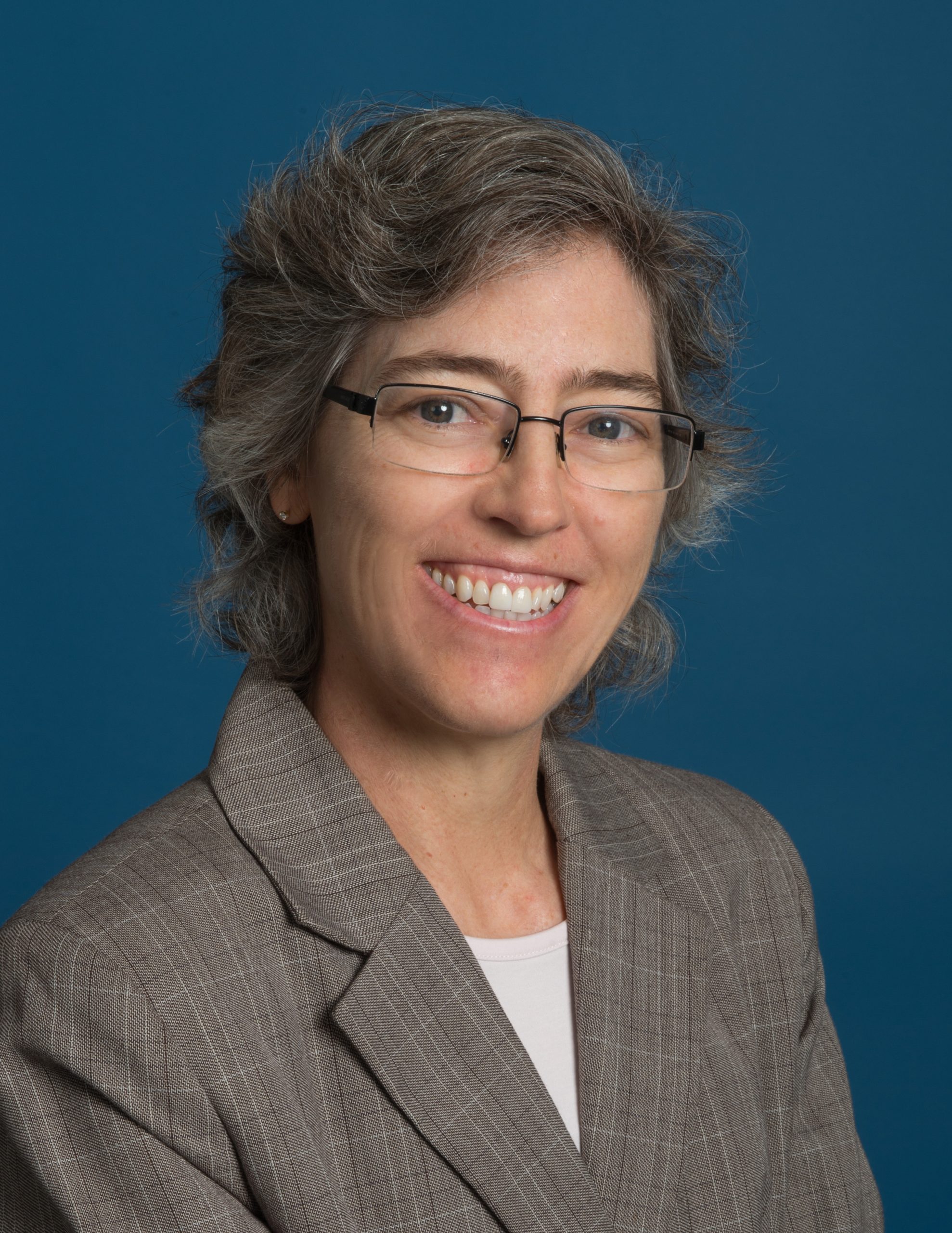 Portrait of Julie Crooke. She is smiling and wearing glasses, a grey blazer, and a white shirt.