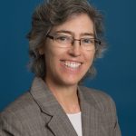 Portrait of Julie Crooke. She is smiling and wearing glasses, a grey blazer, and a white shirt.