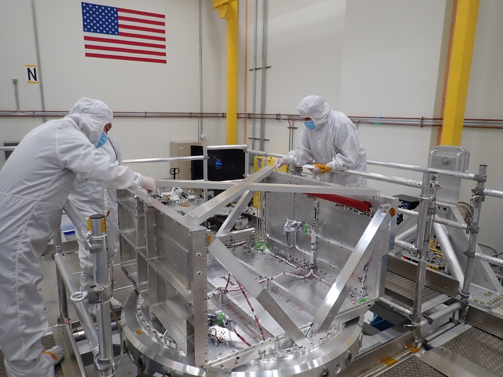 Workers in white protective clothing lean over a big, silver-colored, open ended box that will house and protect Europa Clipper electronics.