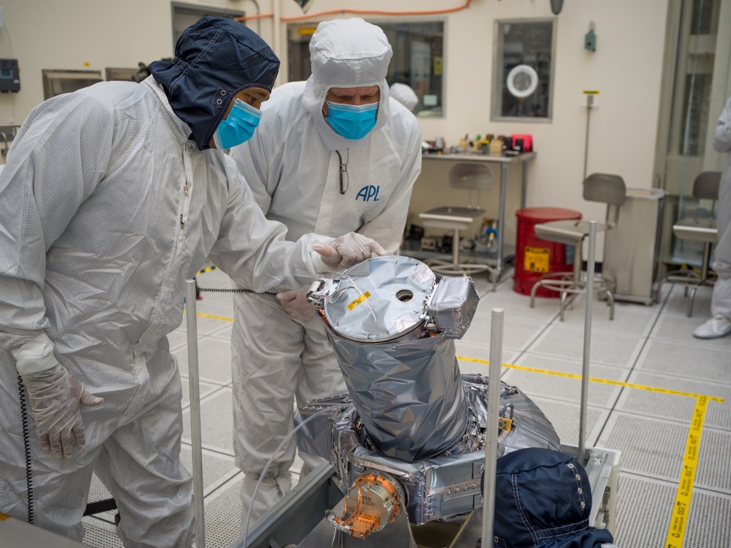 Two workers in protective clothing point and lean toward a piece of equipment covered in silver material.