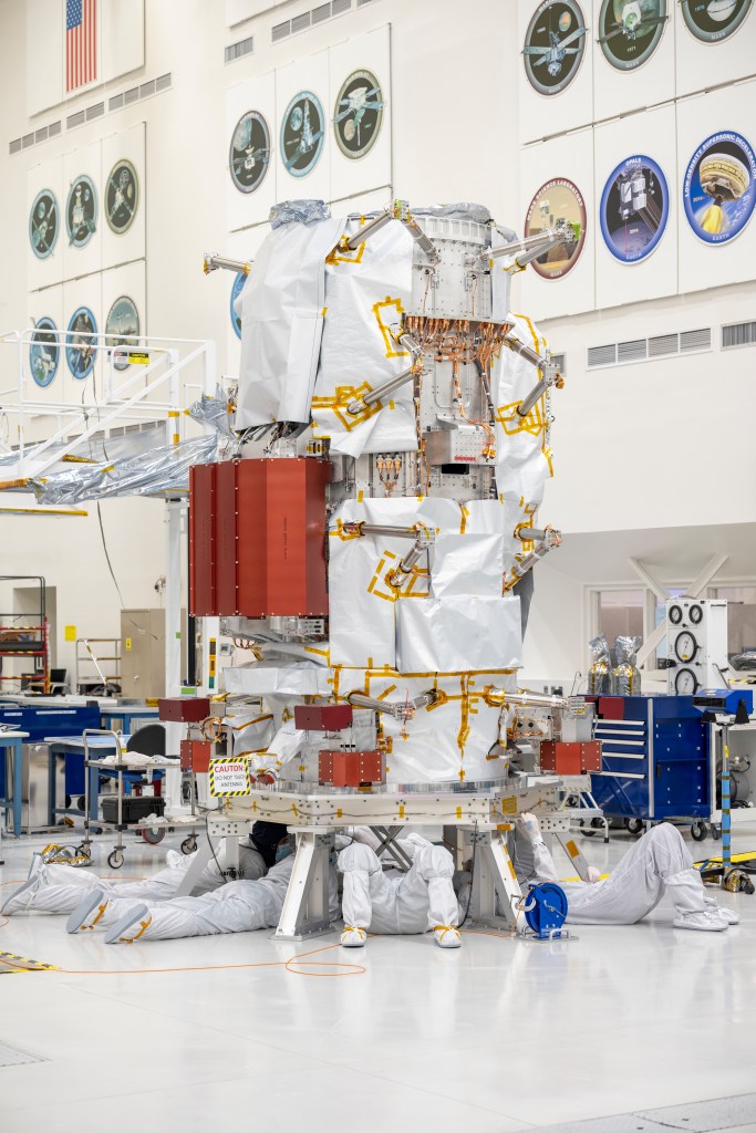 Workers in white protective clothing are on their backs and stomachs working underneath the main body of the Europa Clipper spacecraft. The spacecraft is mostly covered in protective materials but orange-red boxes and copper-colored wires are visible.