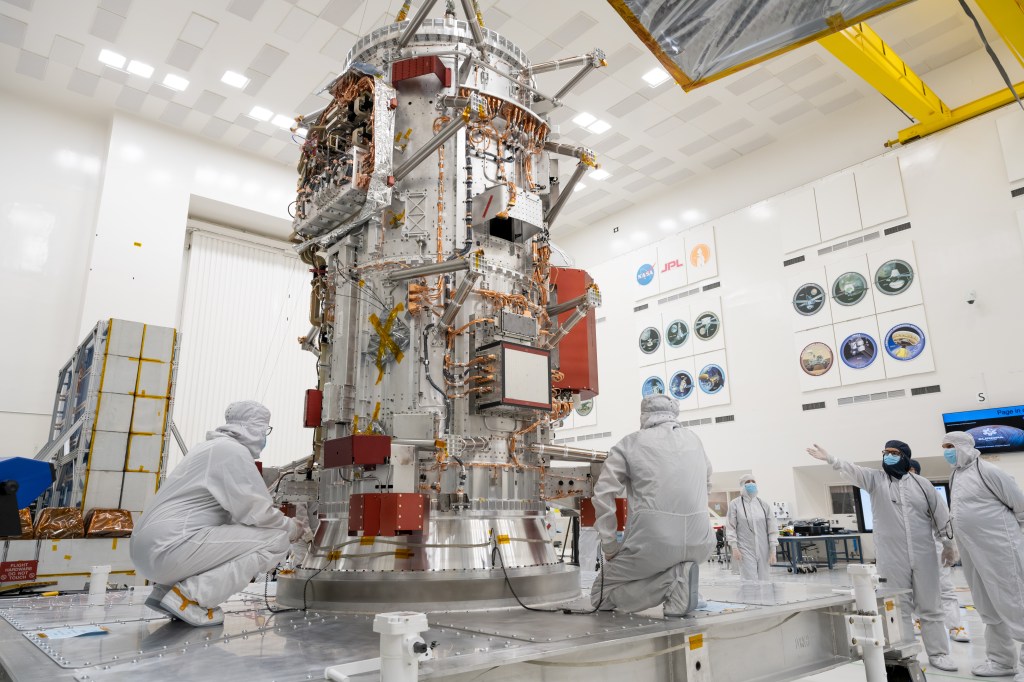 Workers in protective clothing and head covers sit and stand near the main body of Europa Clipper - a tall, silver cylinder with multiple orange and copper cables running between red-orange boxes on the exterior of the cynlinder.