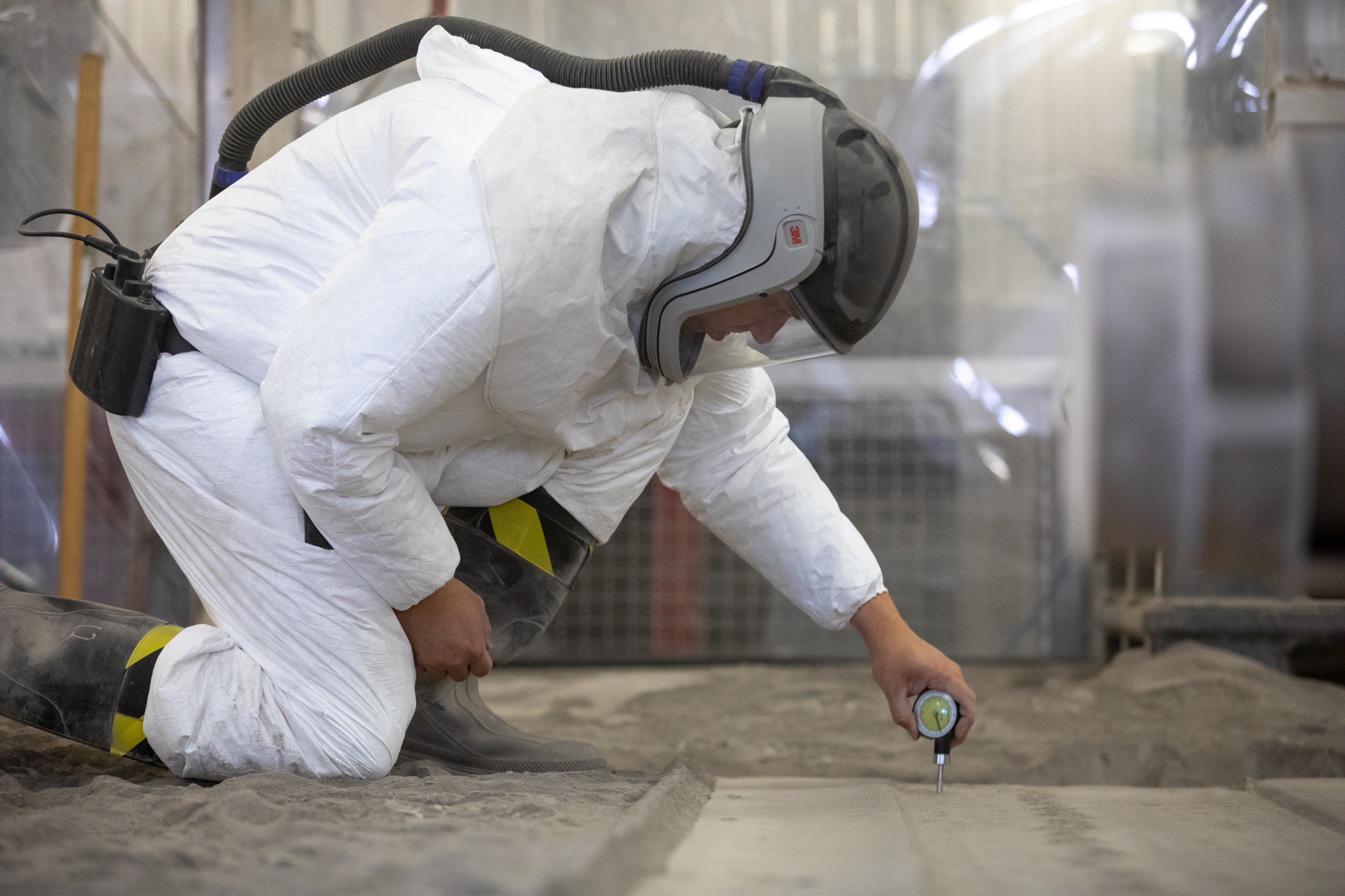A scientist in a safety suit kneels on a bed of simulation regolith and takes measurements from the soil using an instrument that pierces the surface.