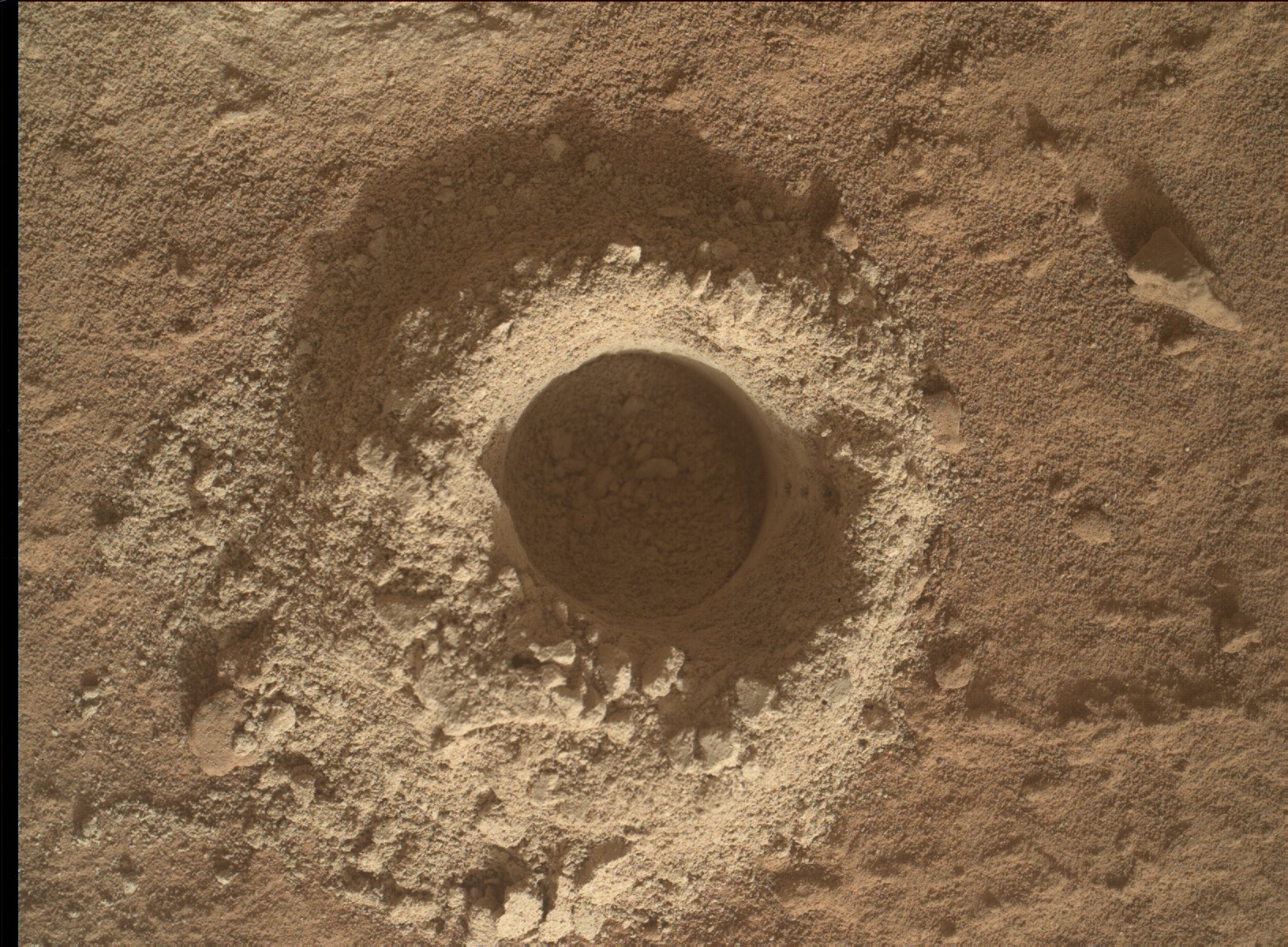 An overhead image of Martian terrain, light orange-brown and resembling a bundt-cake pan viewed from above. A dark drill hole is at center, surrounded by a doughnut-shaped area of soft-looking soil scraped from the hard-packed surface.