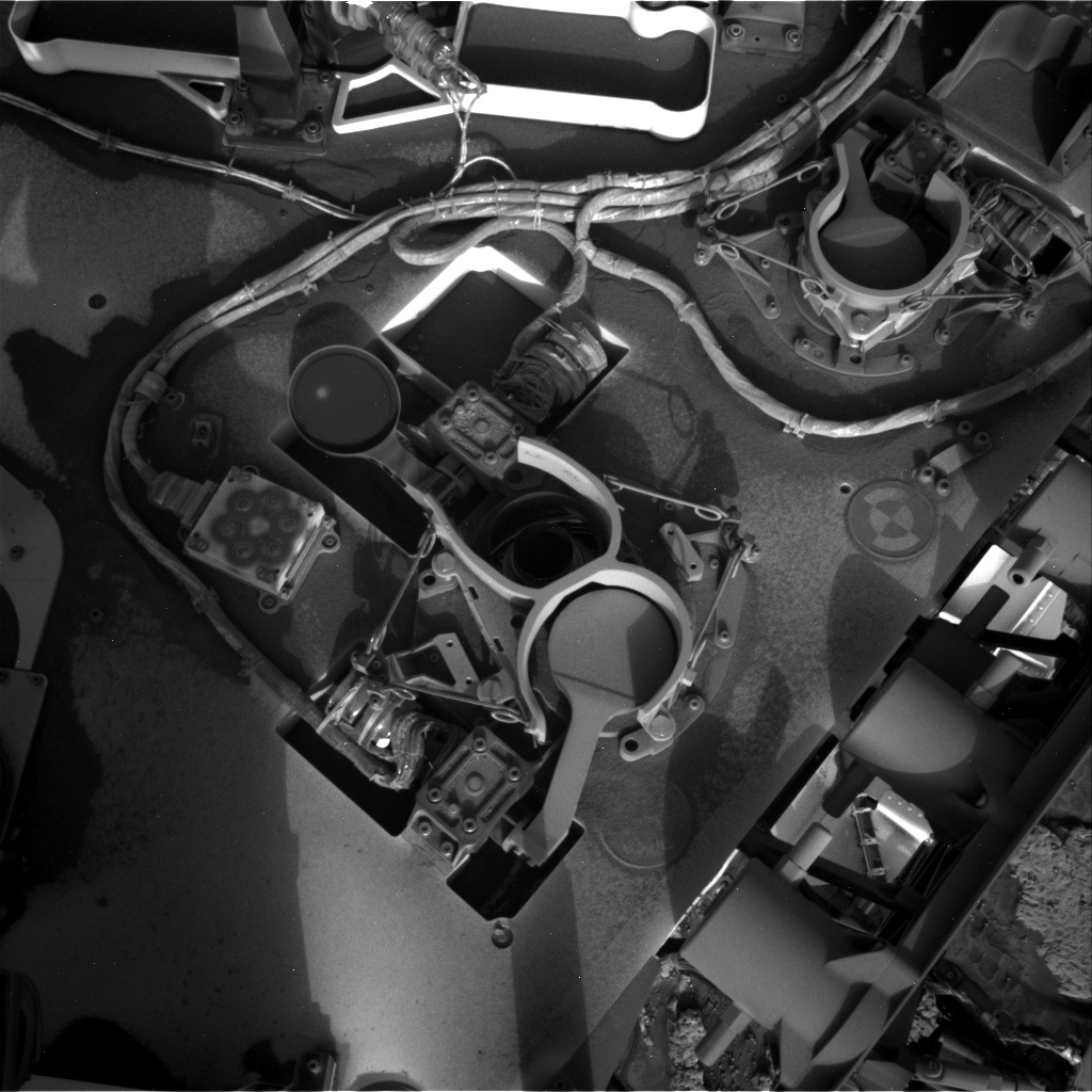 A black and white photograph of a flat, dust-covered section of the Curiosity rover. Cable snake through and around the area shown, which includes three recptacles in different spots, with flip-top caps, each resembling the inlet to refill a car's windshield-wiper fluid. The one at frame center is open, revealing a smaller hole inside.