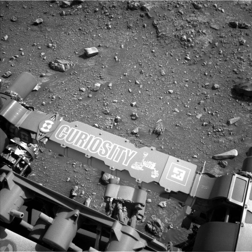 A grayscale photograph of the Martian surface from the Curiosity rover captures parts of the rover in the bottom half of the frame, including a crosspiece imprinted with its name and a line drawing of Curiosity. Ahead of the rover, rough, pebbly terrain in various shades of medium gray fills the rest of the frame, with scattered light gray rocks poking out of the soil. A medium-sized rock at upper left, the focus of the rover's attention, resembles a medium gray pita sandwich sitting upright on the ground.
