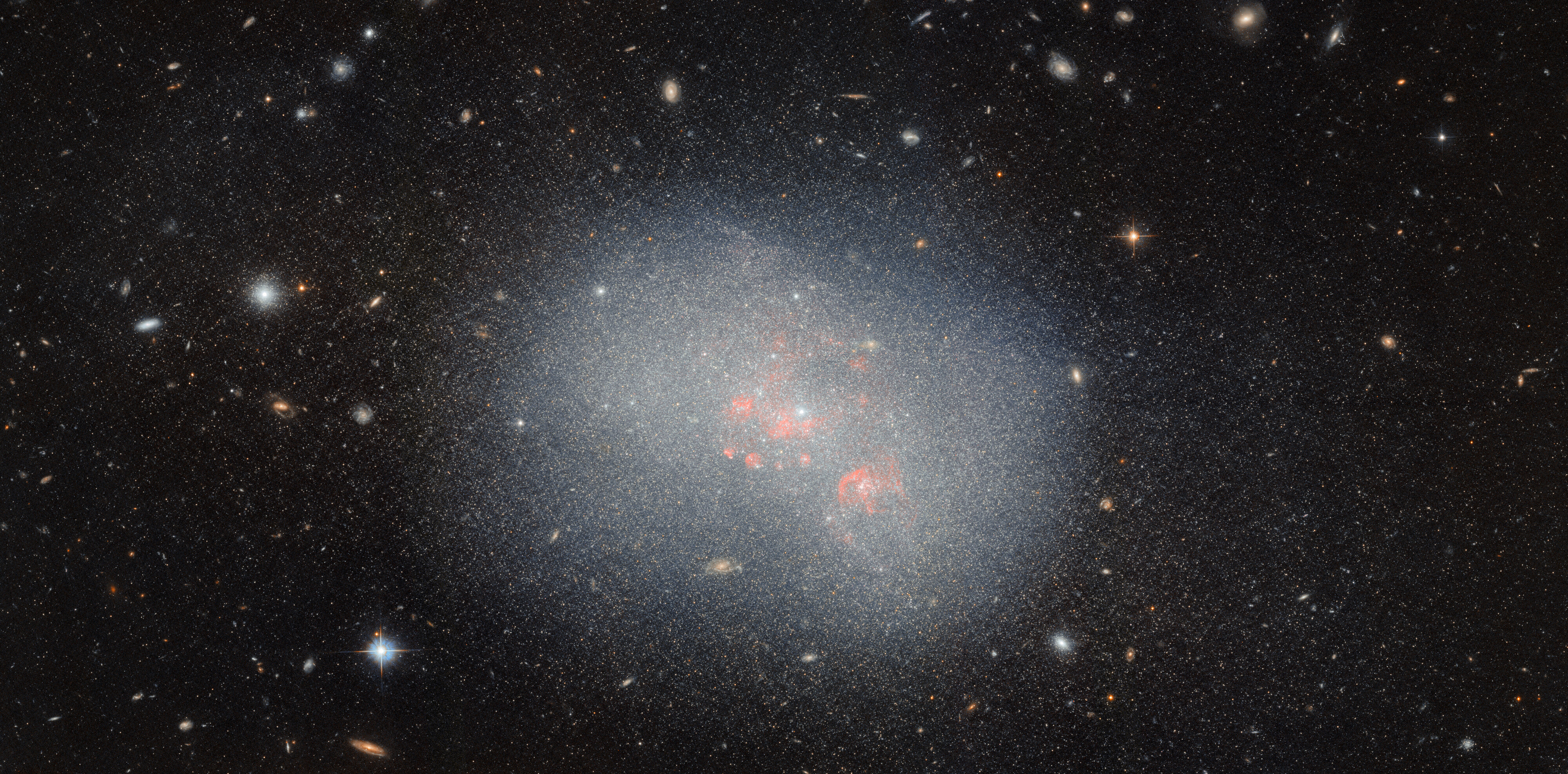A dwarf irregular galaxy. It appears as a cloud of bluish gas, filled with point-like stars that spread beyond the edge of the gas. A few glowing red clouds sit near its center. Many other objects are visible around it: distant galaxies in the background, four-pointed stars in the foreground, and star clusters that are part of the galaxy appear as bright spots surrounded by more tiny stars.