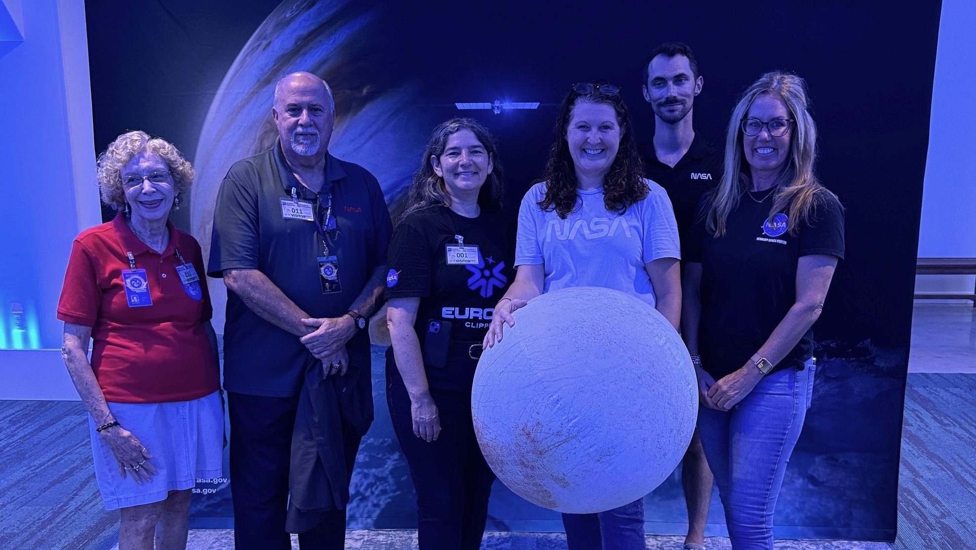 Six people working at the Europa Clipper Roadshow in New Orleans pose in front of a poster of Jupiter, Europa and the Europa Clipper spacecraft. One person holds a beachball designed to look like Europa.