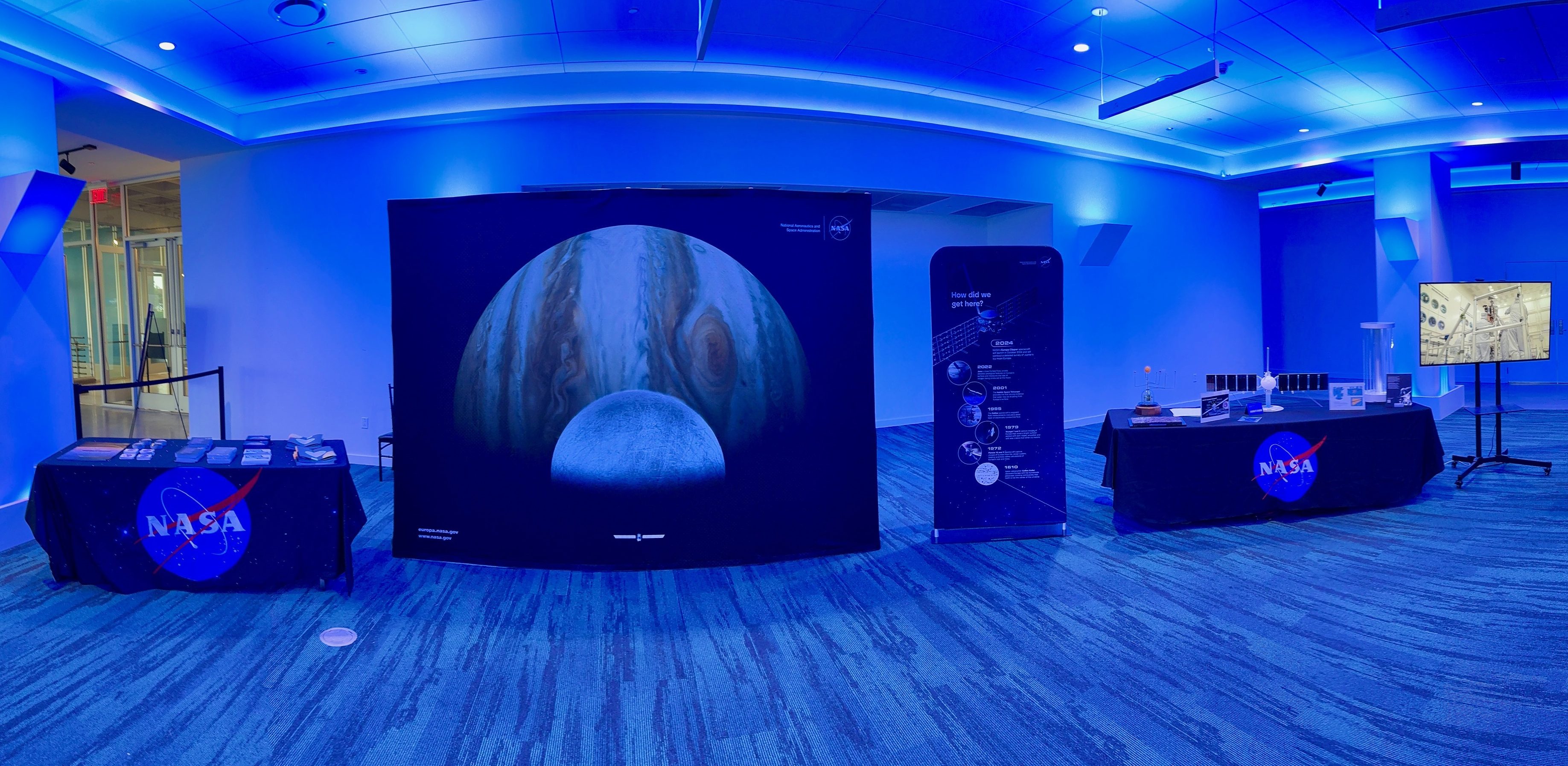 An exhibit that includes multiple tables with black tablecloths with the NASA logo, a selfie station with a large image of Jupiter, Europa, and Europa Clipper; and a large monitors.