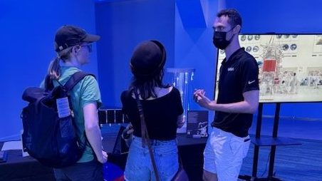 Two people talk to a NASA/JPL public engagement specialist in front a table with a monitor off to one side.