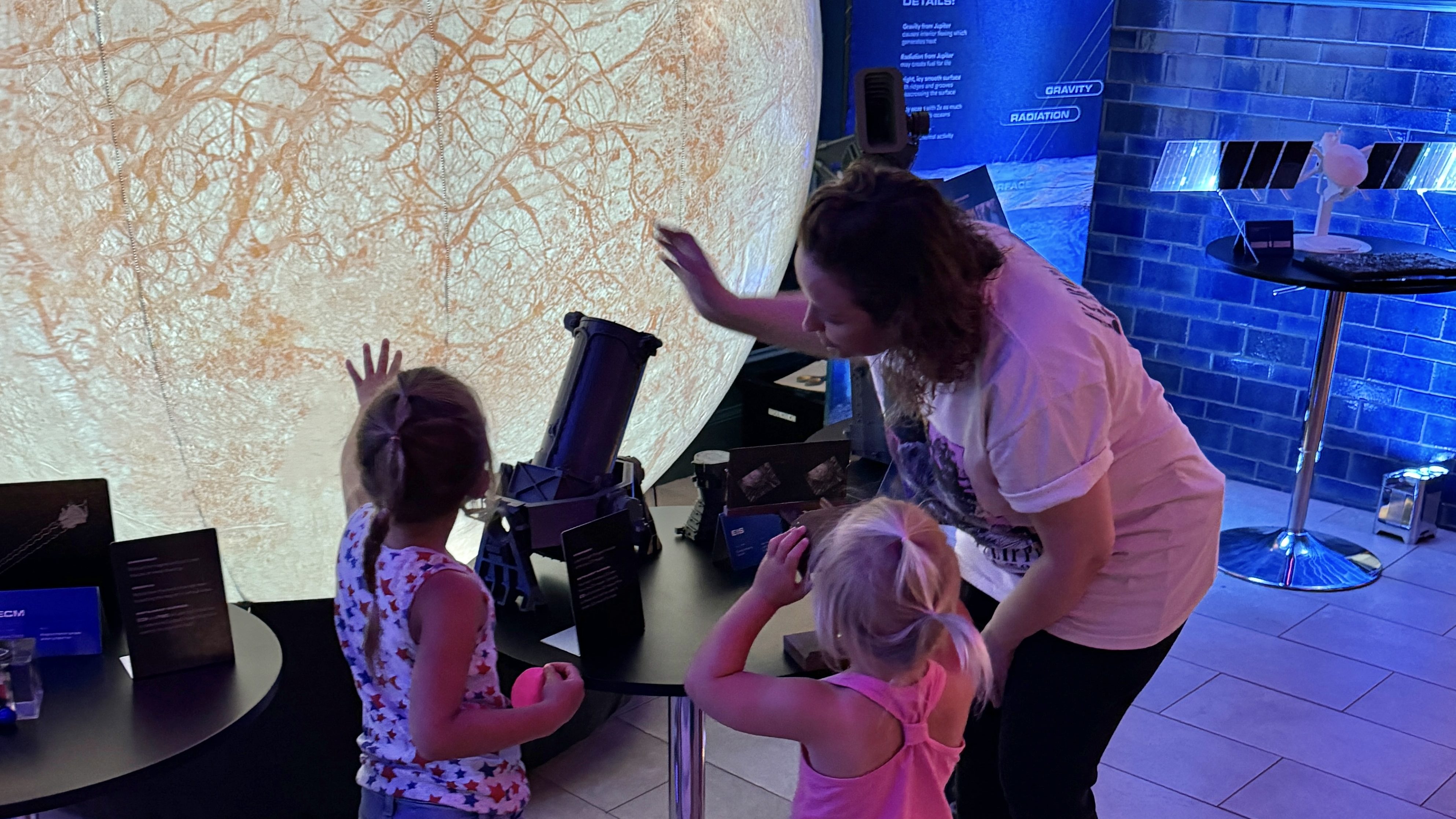 Two young children stand at a round exhibit table in front of an inflatable model of Europa as a woman leans down to explain items in the exhibit.