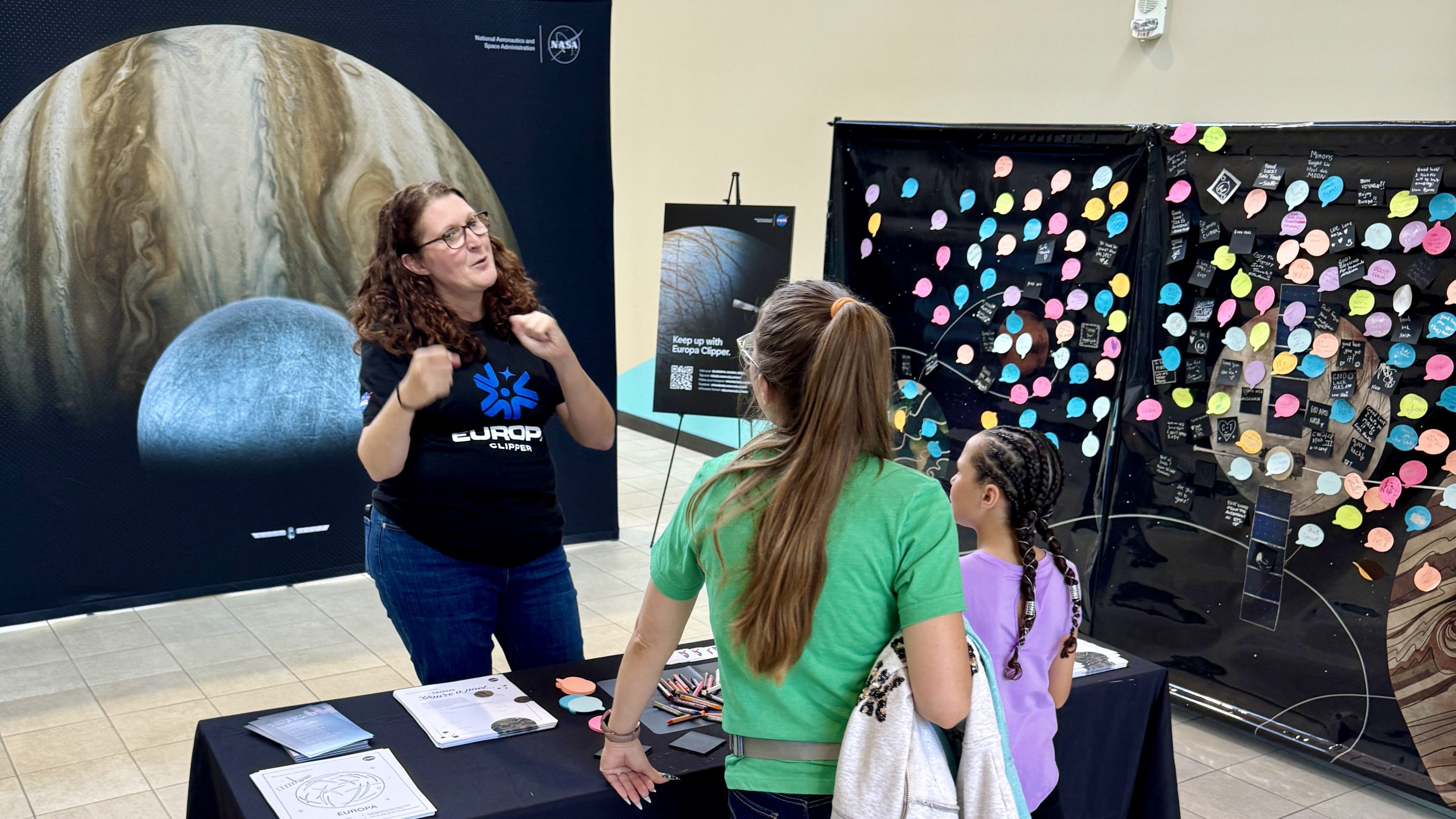 A member of NASA/JPL's public engagement team stand behind a table and talks to people about the exhibit. Off to one side is a board used to gather notes from the public to members of the mission team. In the back is a Europa Clipper selfie station. It has a large picture of Jupiter with Europa and Europa Clipper in front.