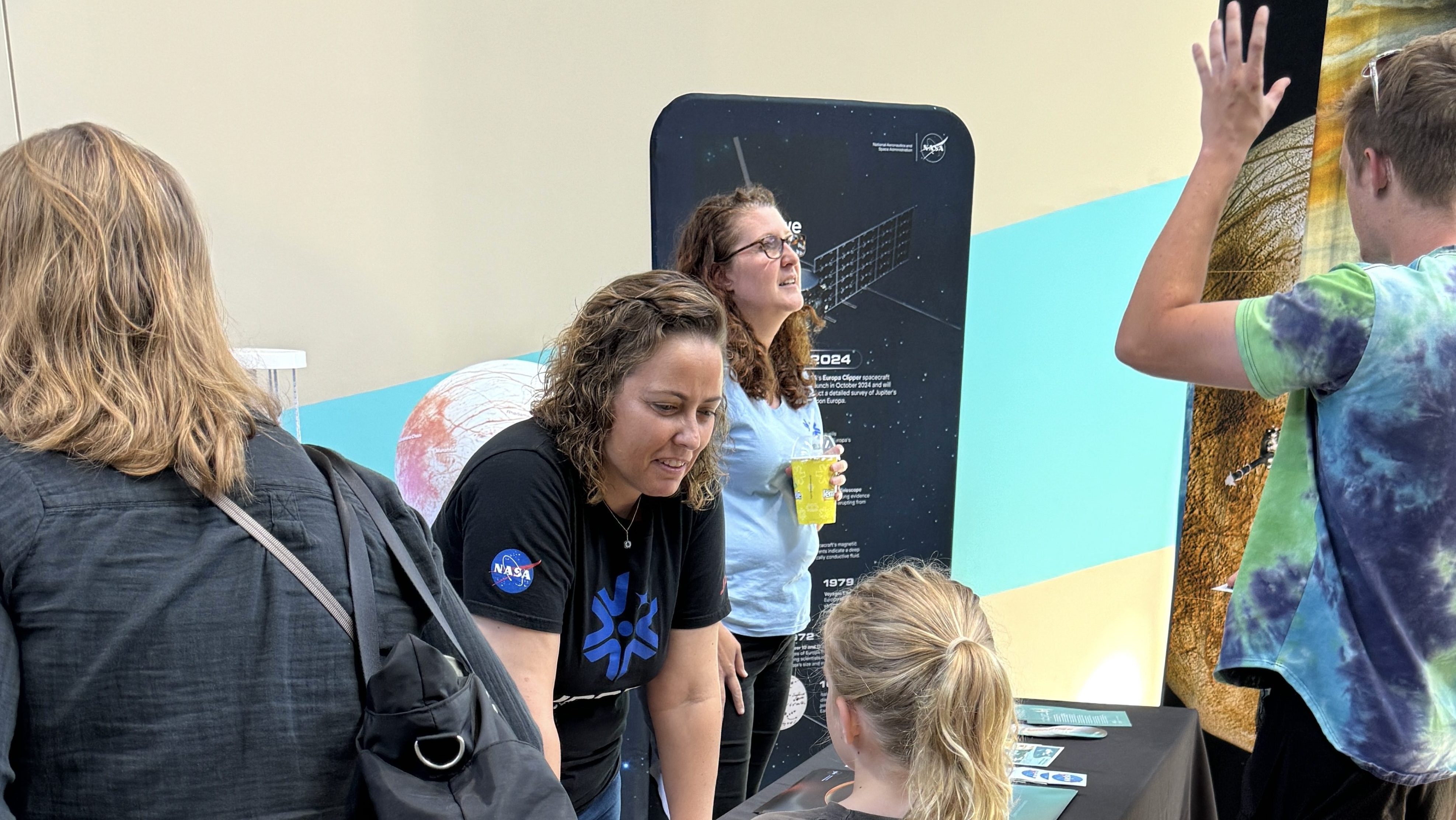 A young child and an adult talk to members of NASA/JPL's solar system public engagement team who are standing behind a table. Another adult looks on. Handouts are on the table, which is covered in a black tablecloth with a NASA logo in the center. Posters can be seen behind the table.