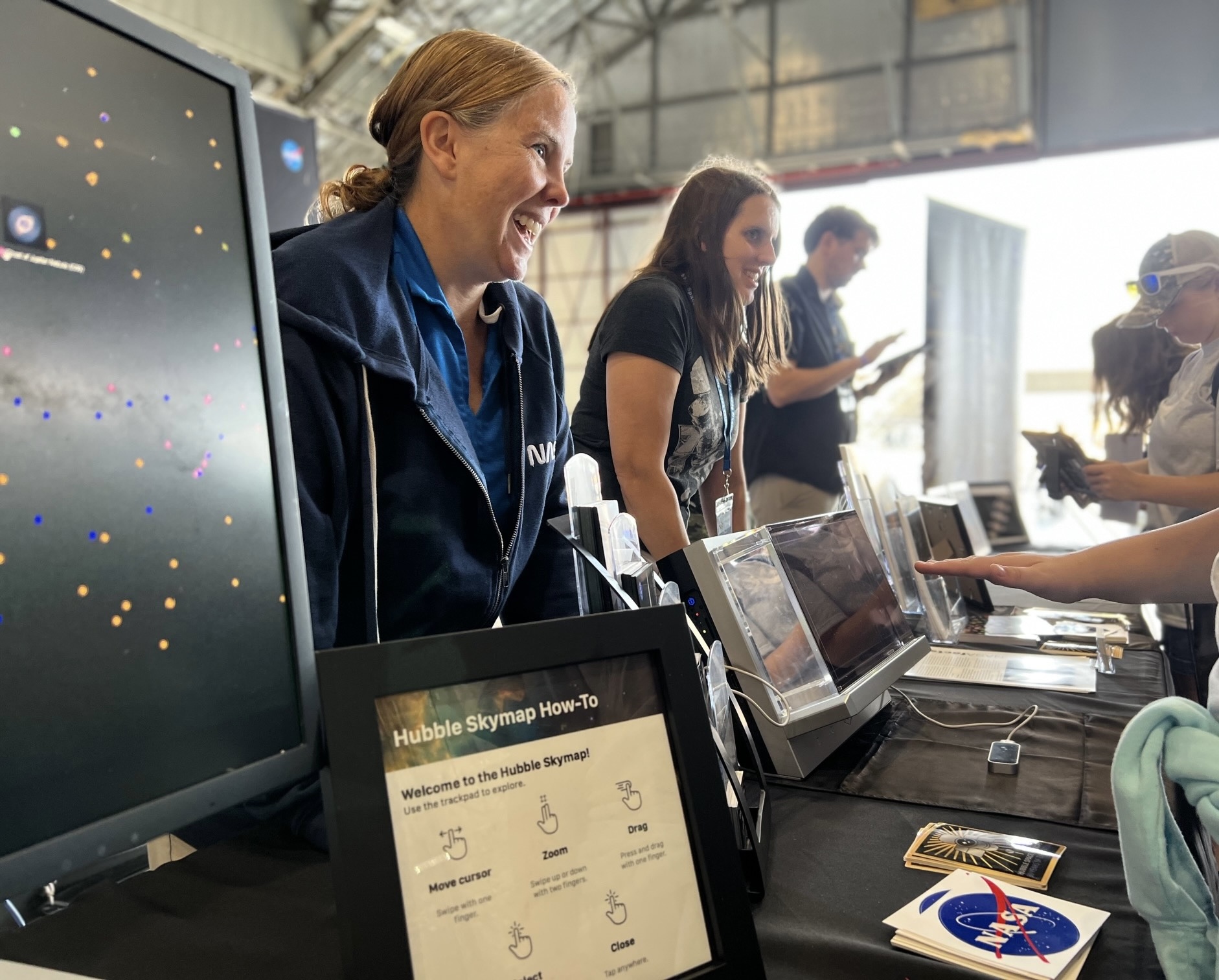 Two women and a man standing at a table with a computer monitor and a digital interactive hubble space telescope speaking to people at an event.