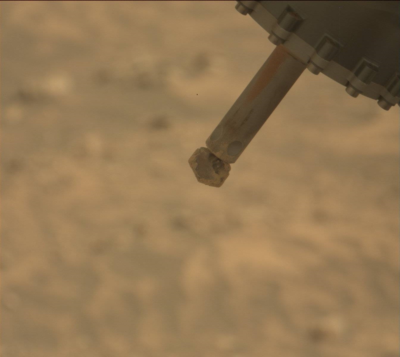 Against a blurry orange-bown backdrop of the Martian surface, an instrument pokes into the frame from upper right. What looks like a gray metal base fills the upper right corner, and extending from that is a spindle, with what looks like a grinding drill bit at its end, terminating at the center of the image.