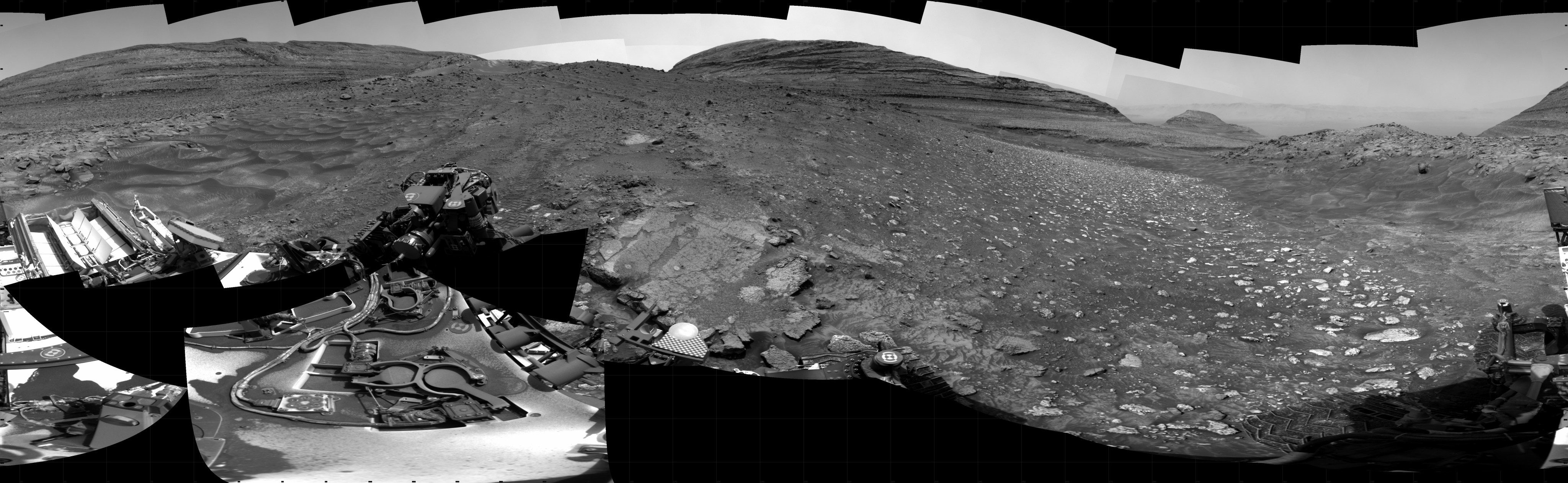 A panoramic black and white image of the Martian landscape before the Curiosity rover. Wind-sculpted, wavy sand dunes appear at far left, while fields of scattered jagged and broken rocks cover the rest of the foreground, while the background is dominated by three large buttes of layered rock.