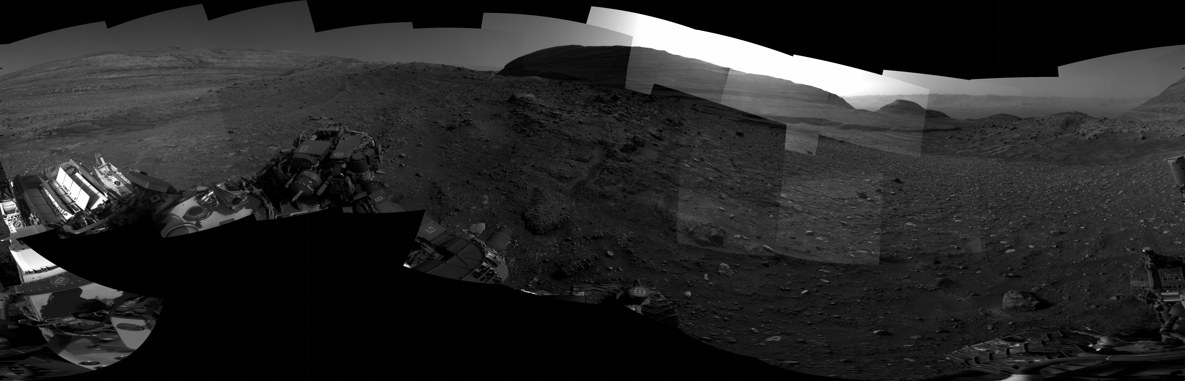 A black and white photographic panorama of the surface of Mars, showing rolling hills in the distance, with the foreground a mixture of areas with soil, gravel and small rocks, of windswept dunes, and of large broken slabs of flat rock.
