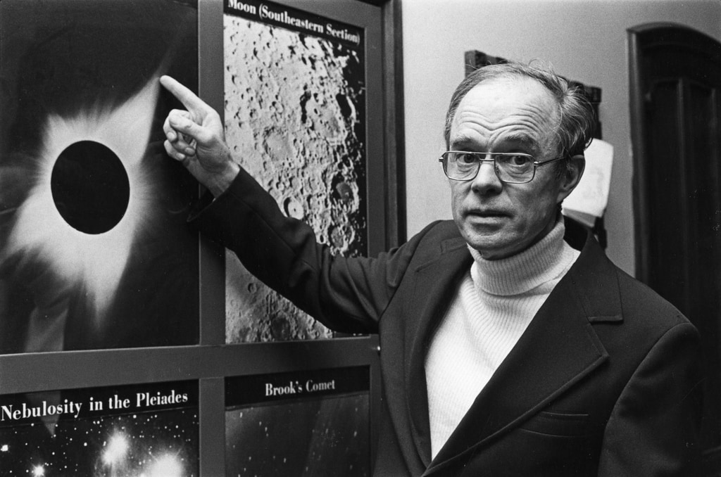 A black and white photograph of a man standing in front of an image of a total solar eclipse. He is pointing at the image.