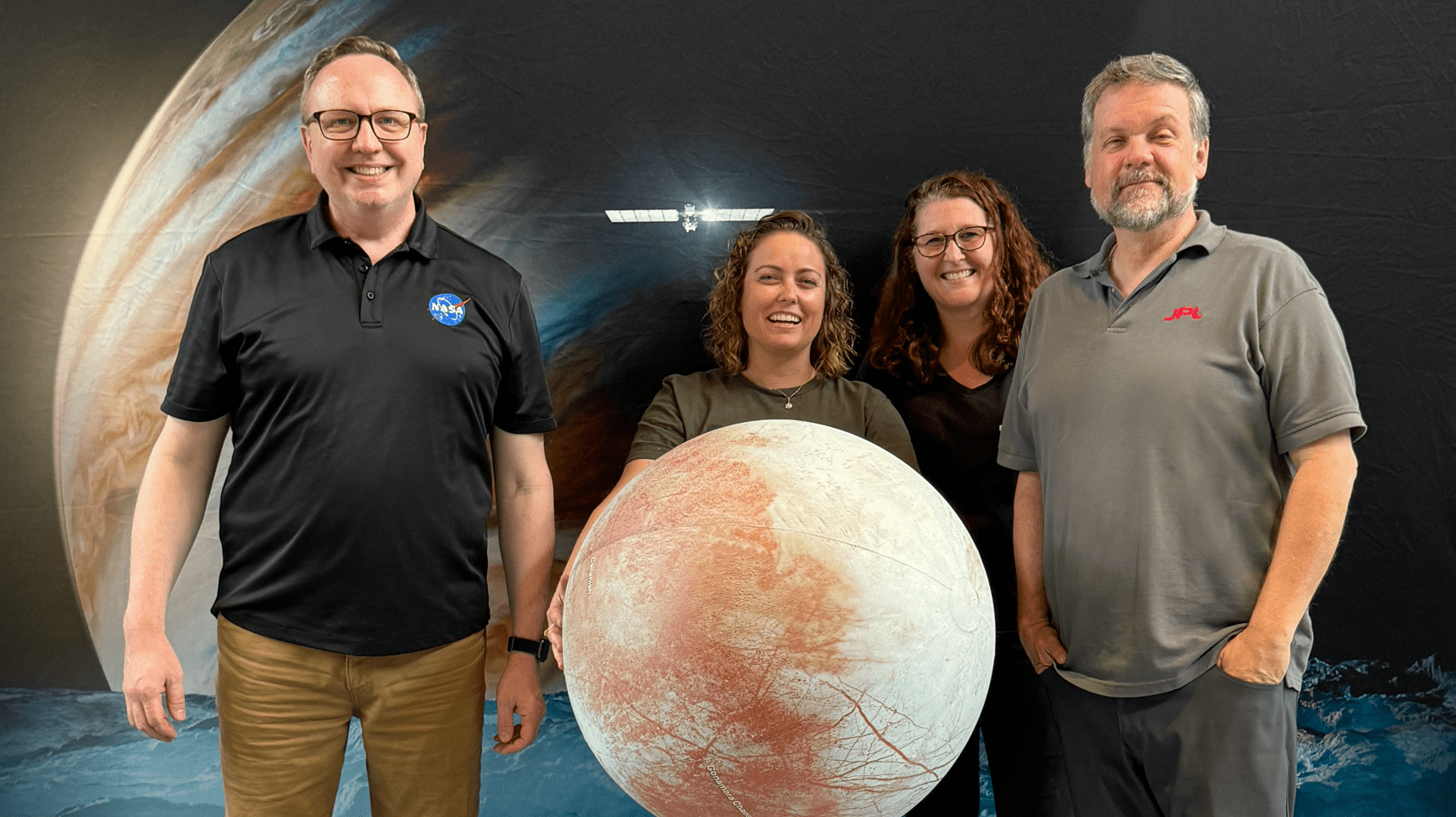 Four members of the Europa Clipper public engagement team pose in front a giant poster showing Jupiter, the Europa Clipper spacecraft, and the surface of Europa.