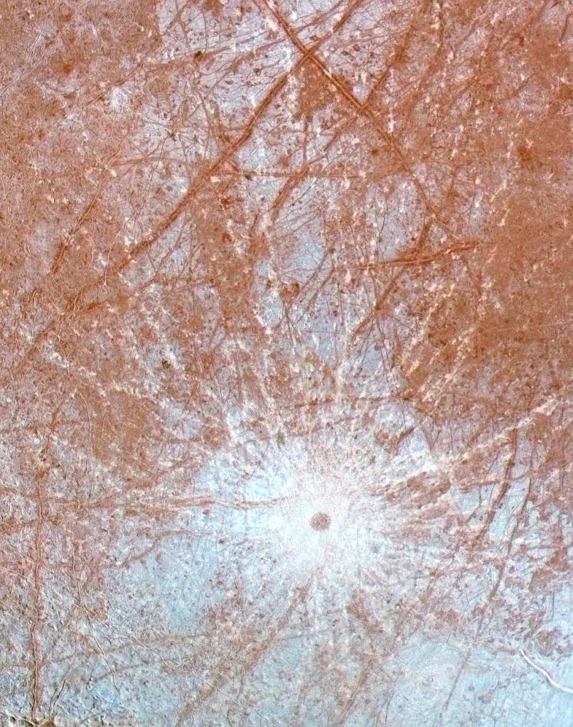 Europa's Pwyll Crater looks like a bright, white circle surrounded by reddish lines criss crossing the moon's surface.